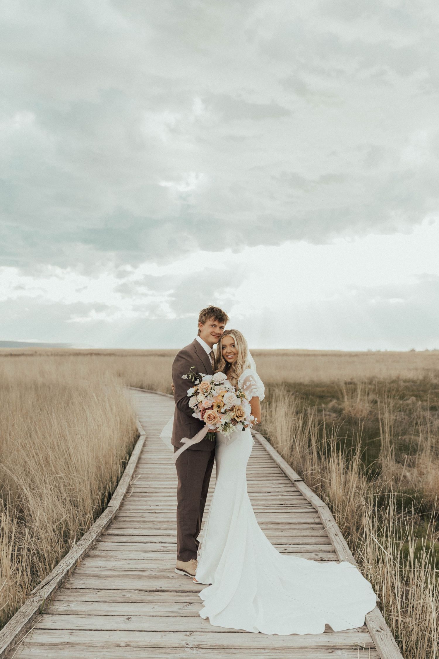 A couple shares a romantic moment on a wooden boardwalk surrounded by tall grass under an overcast sky.