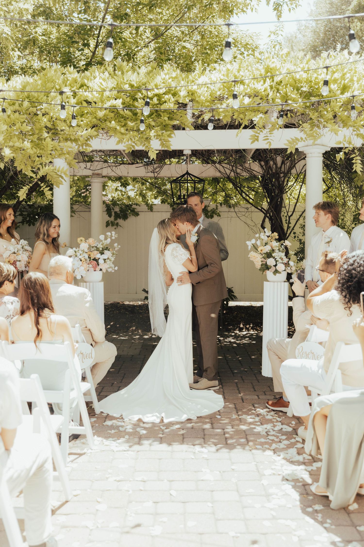 A romantic outdoor wedding ceremony under a floral-adorned gazebo with white chairs and natural sunlight filtering through trees.
