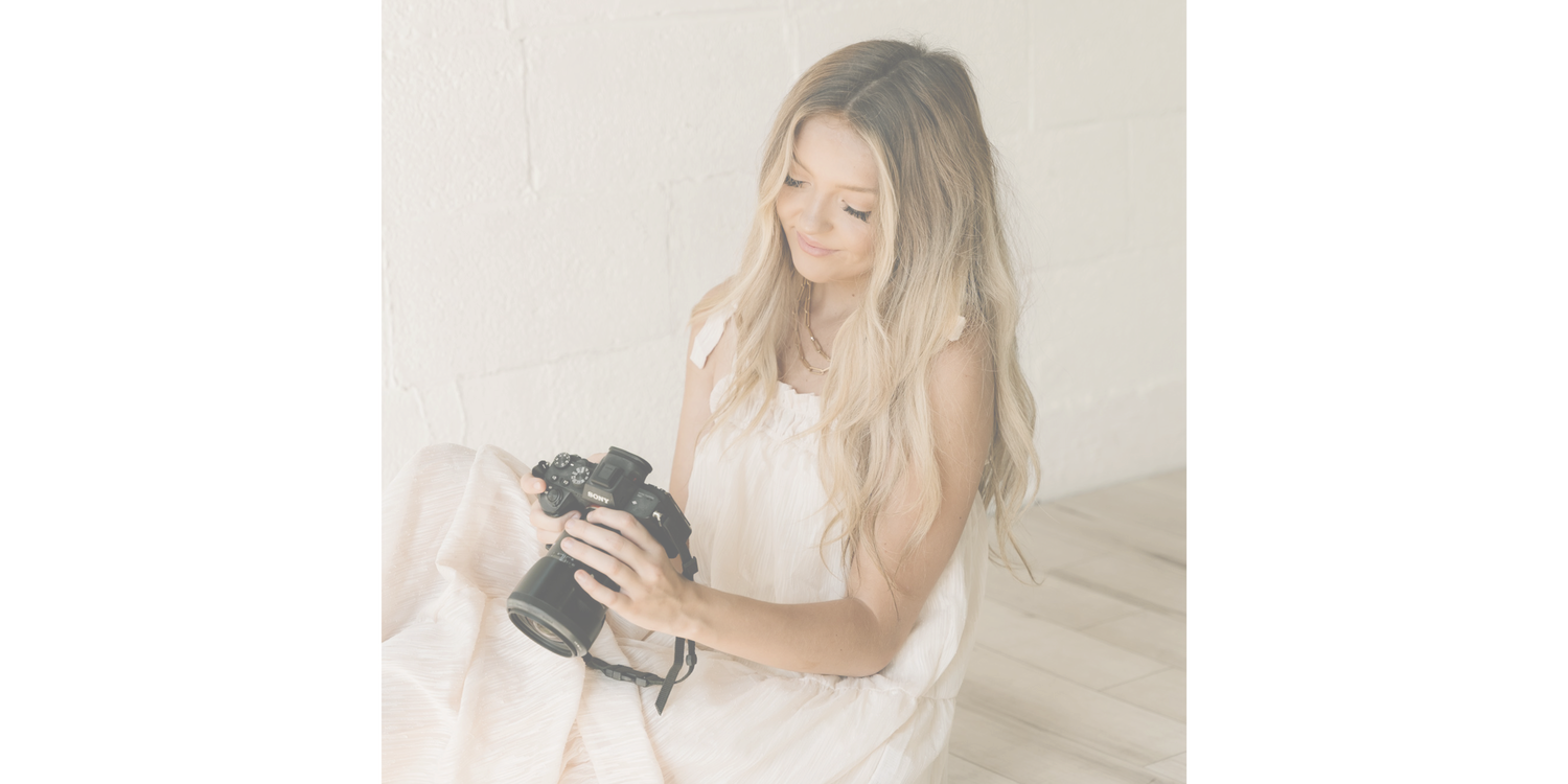 Someone in a white dress examines a camera while sitting in a minimalist neutral-toned setting.