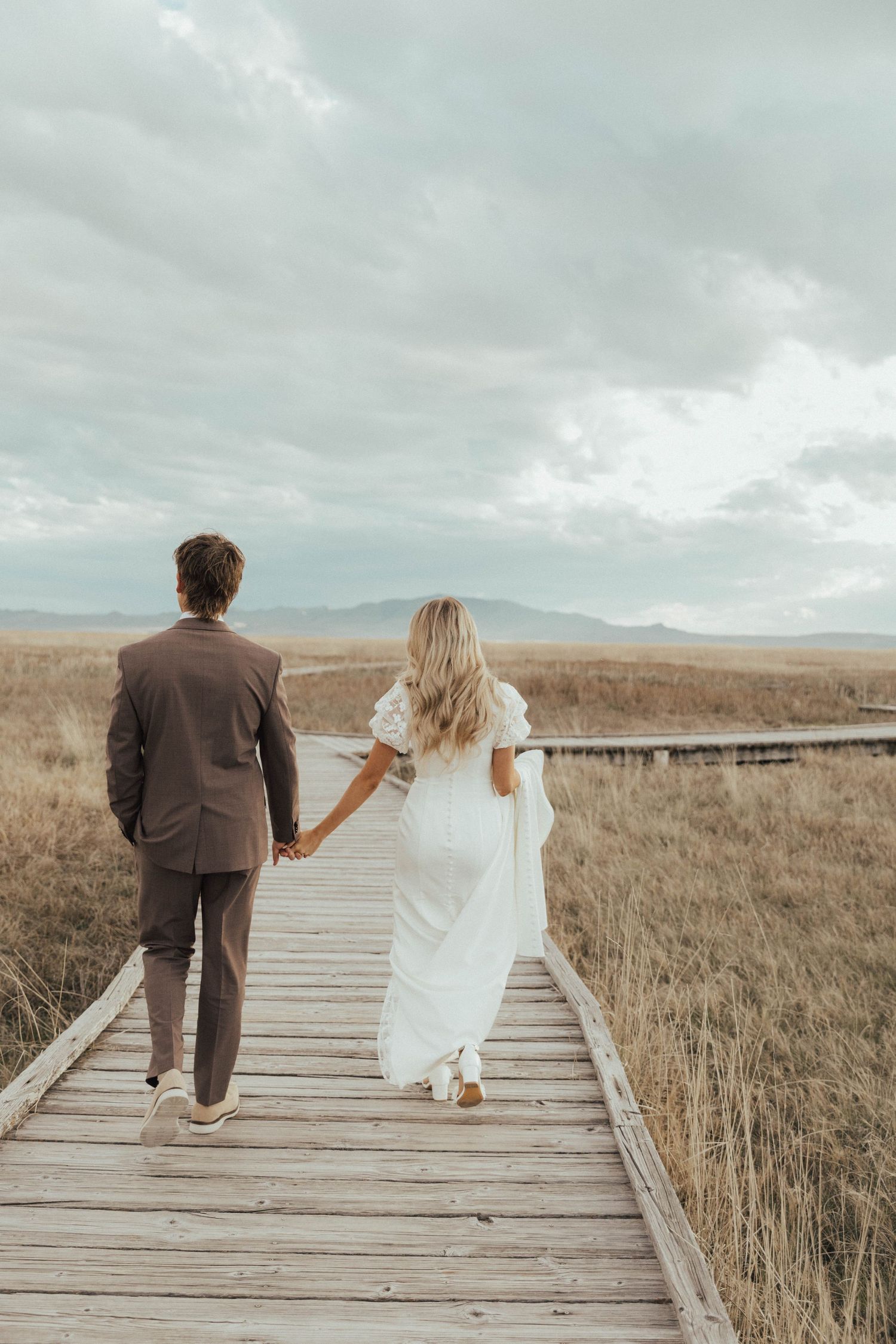 A couple walks hand in hand on a wooden boardwalk through a grassy field beneath cloudy mountain skies.