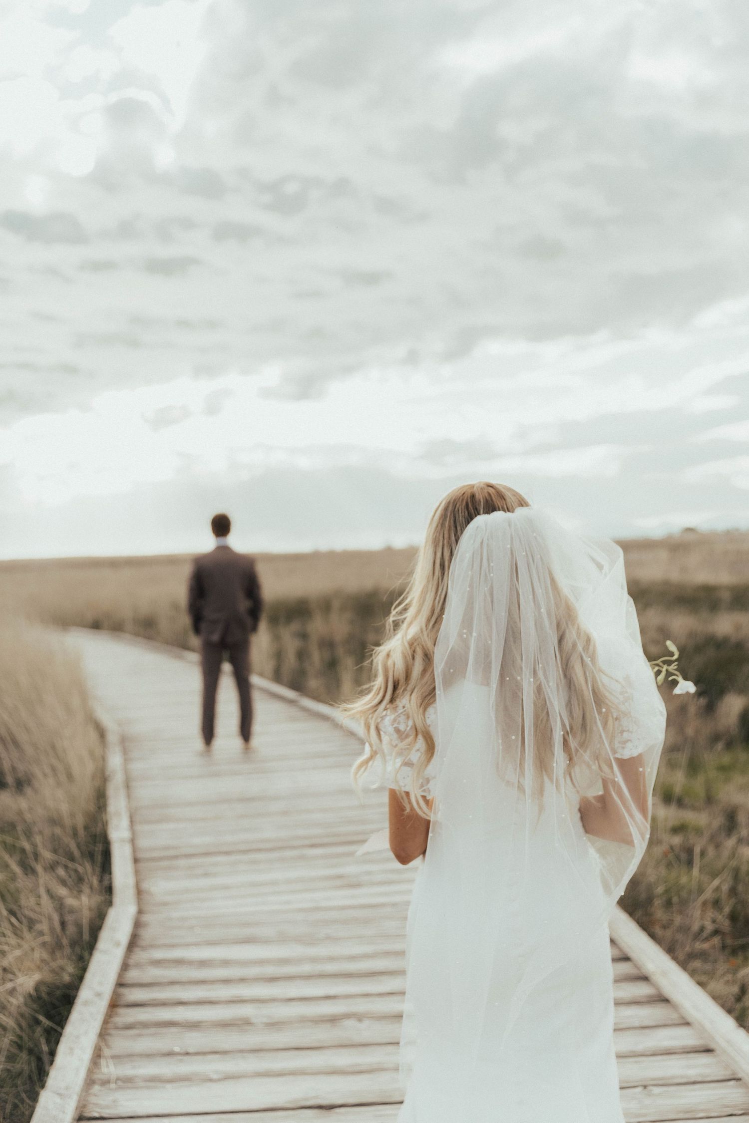Bride in flowing white dress and veil walks along wooden boardwalk toward groom in moody outdoor setting.