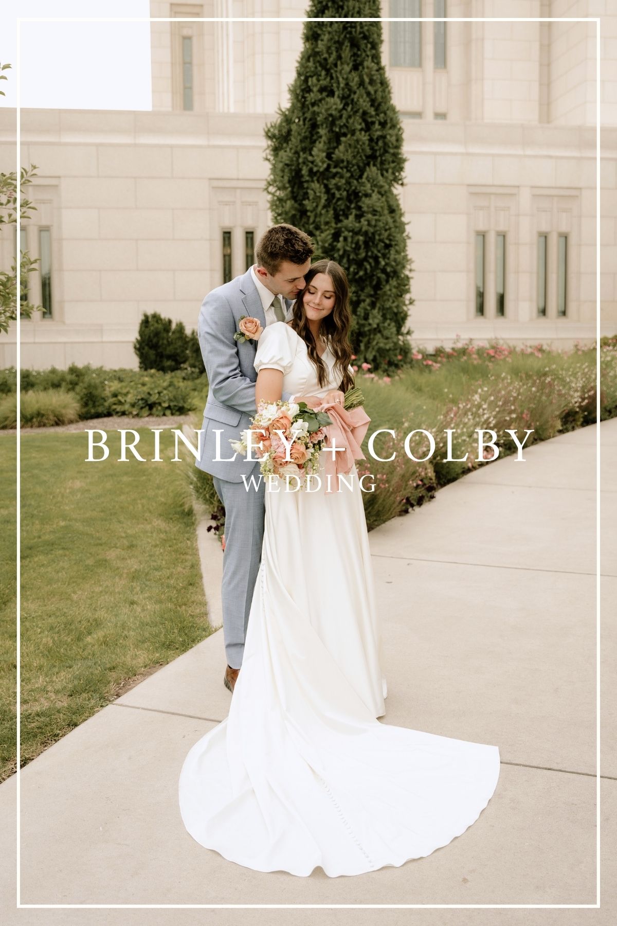 A couple in wedding attire embrace on a landscaped walkway next to a tall cypress tree and light-colored building.