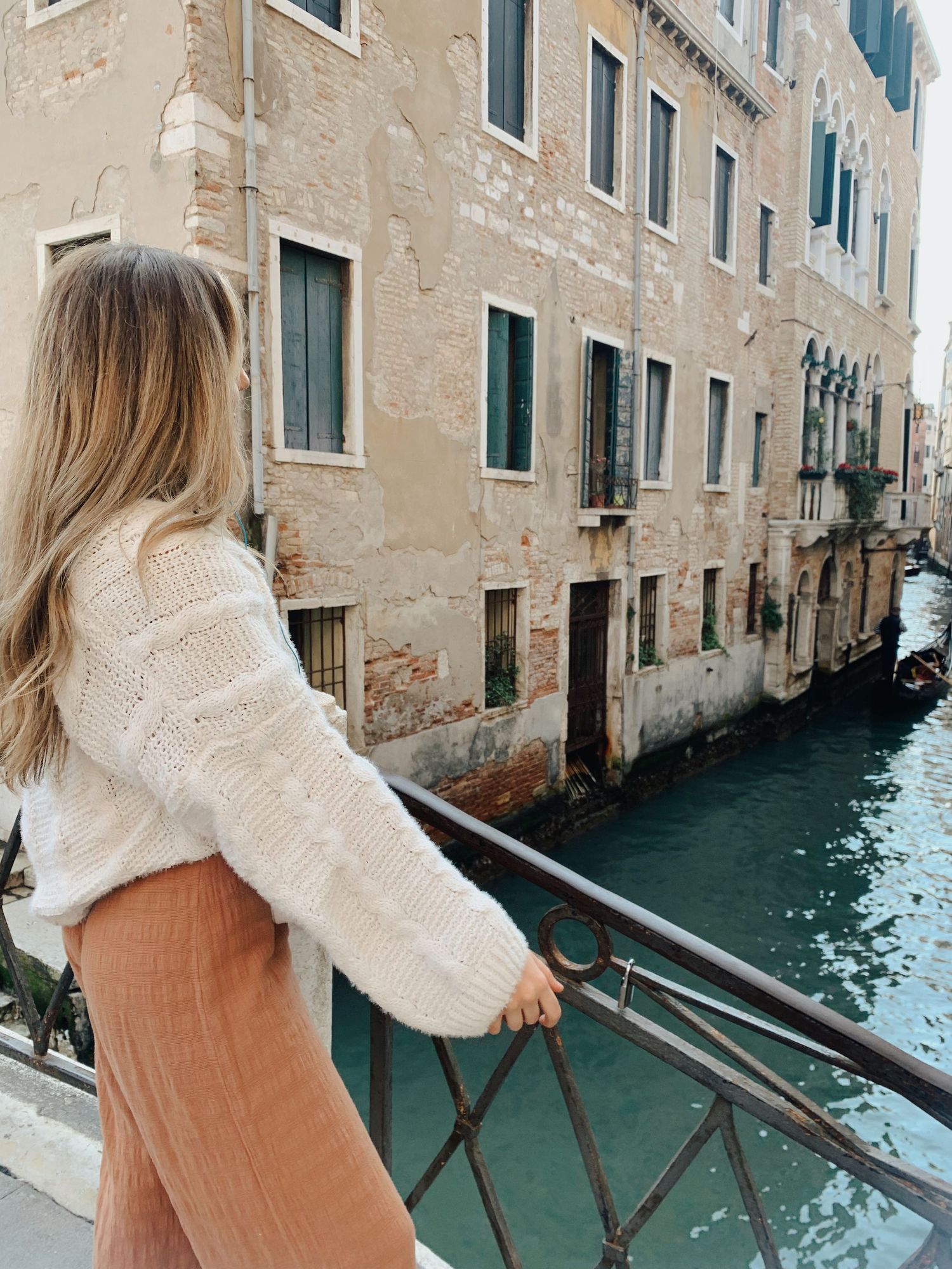 Tourist in cozy white sweater admires historic Venice canal from bridge railing against weathered beige buildings.