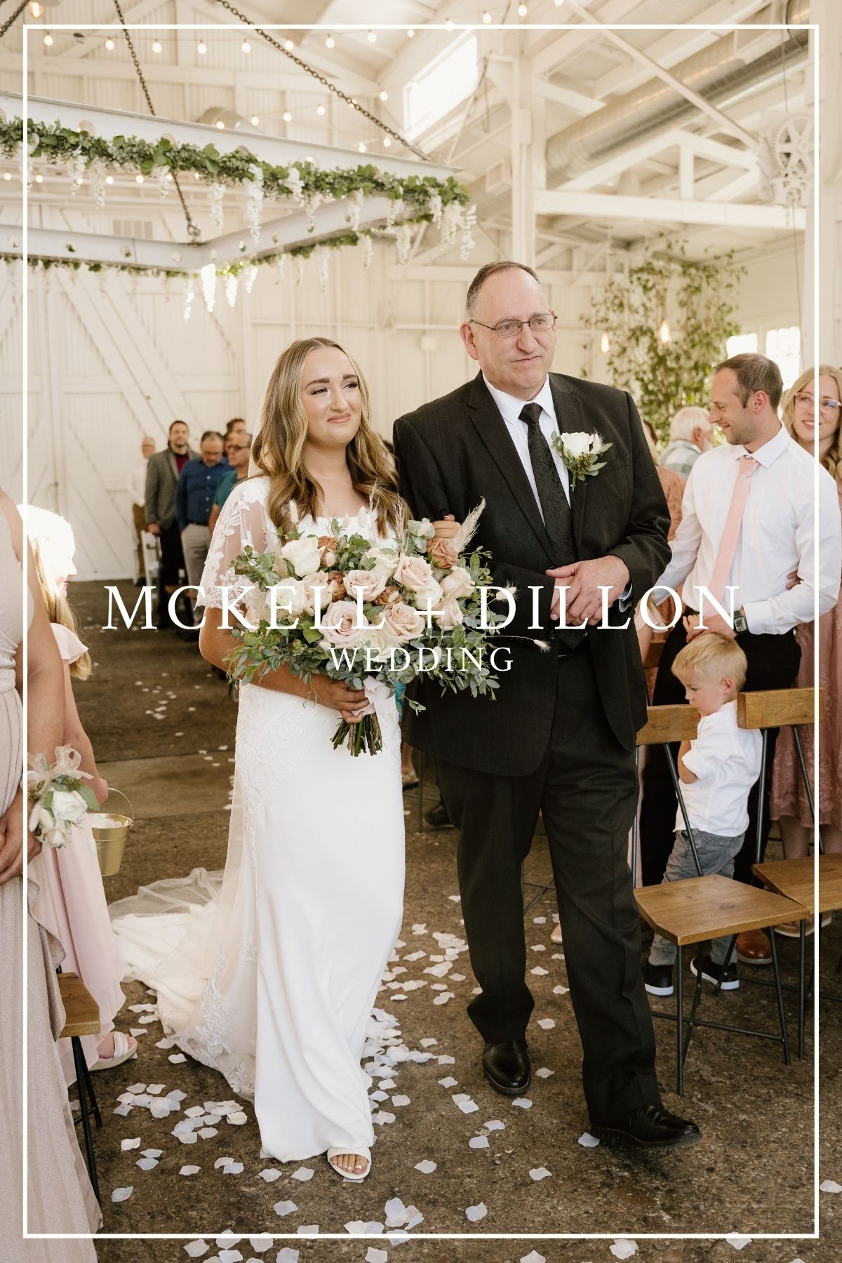 A wedding ceremony processional in a bright indoor venue decorated with greenery and flower petals along the aisle.