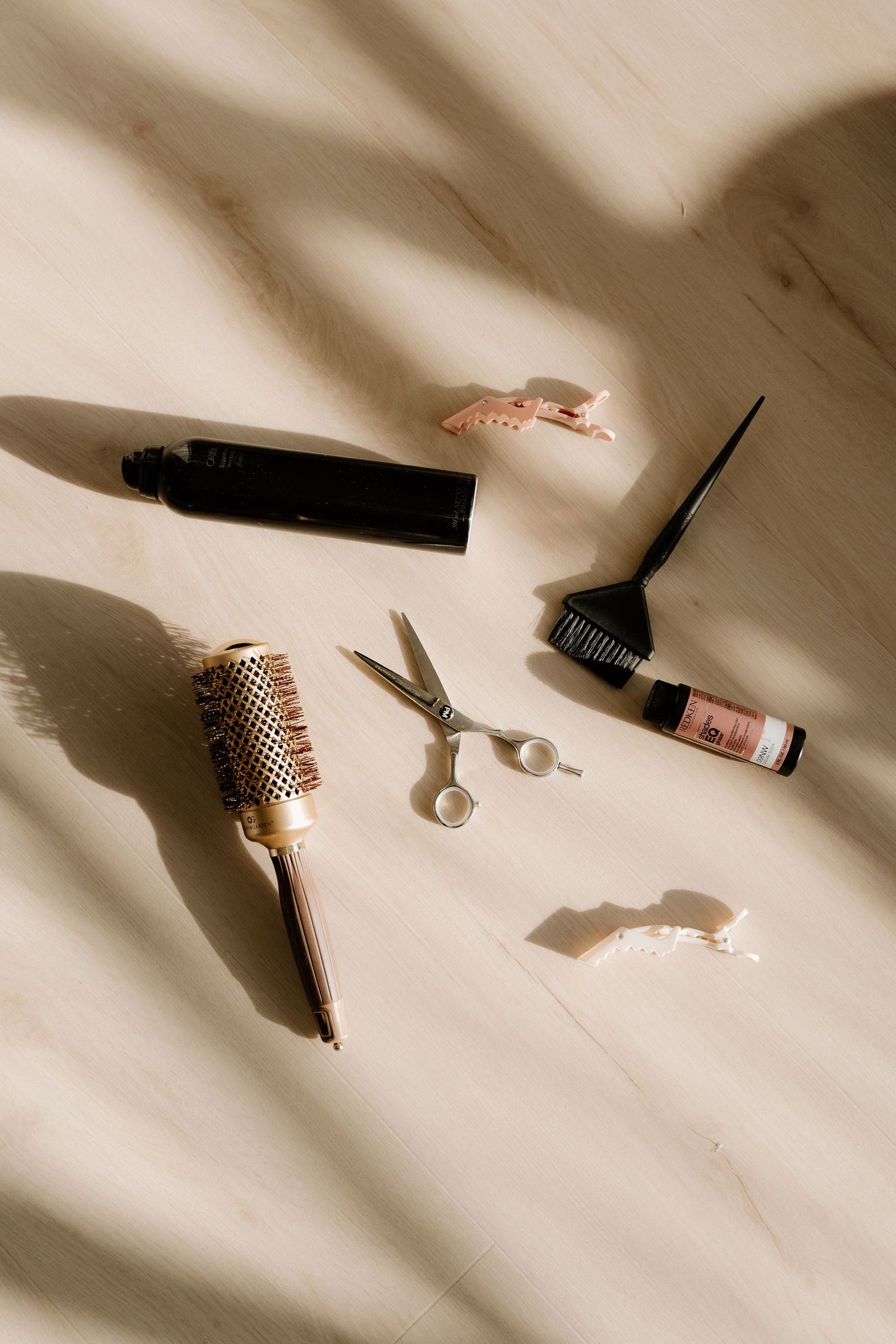 Hair styling tools including round brush, scissors, and beauty products arranged on beige fabric with dramatic shadows.