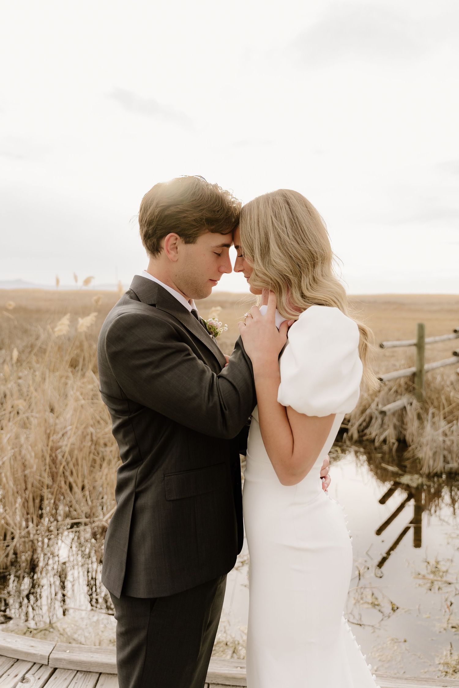 A romantic couple shares an intimate moment on a wooden boardwalk surrounded by tall grass and overcast skies.