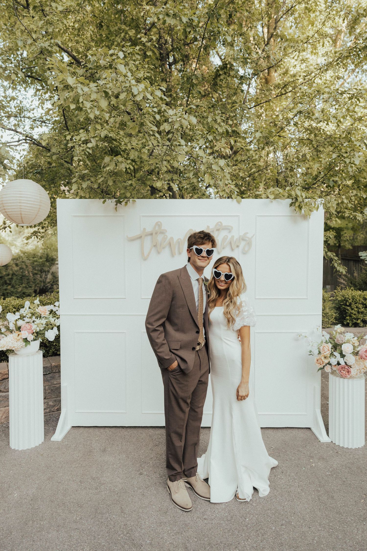 A couple poses against a white backdrop decorated with flowers at an outdoor wedding ceremony.