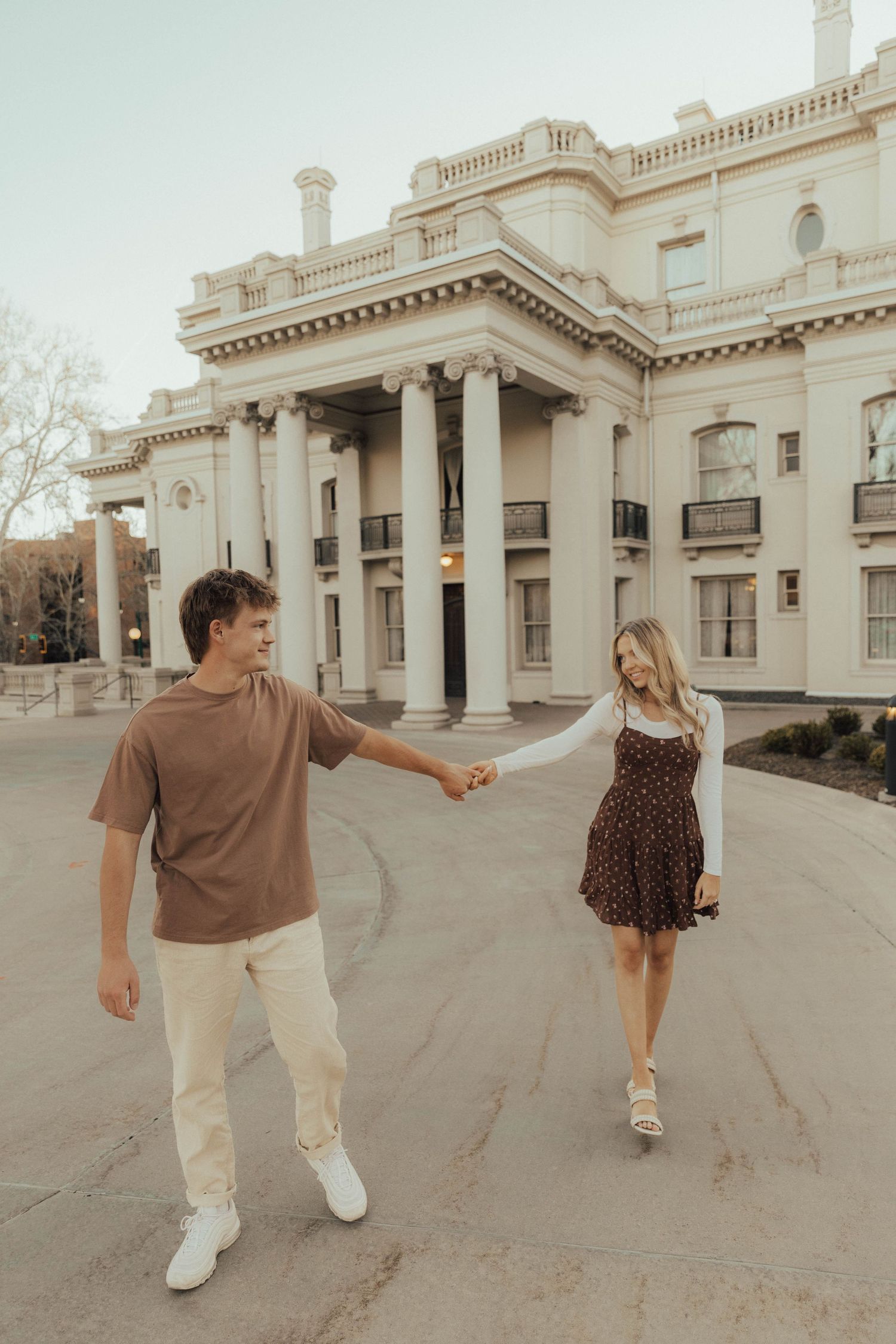 A couple holds hands while walking along a path in front of a grand historic mansion with white columns and architecture.