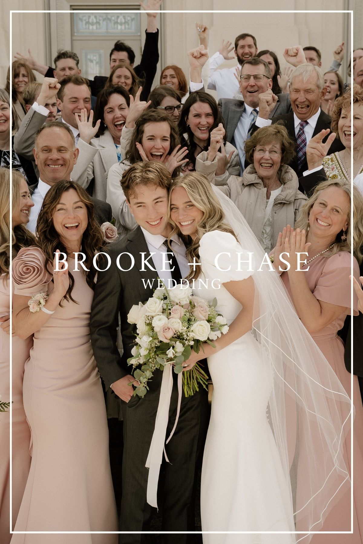 A joyful wedding group photo with the bride in white holding a cream floral bouquet, surrounded by guests in blush and gray tones.