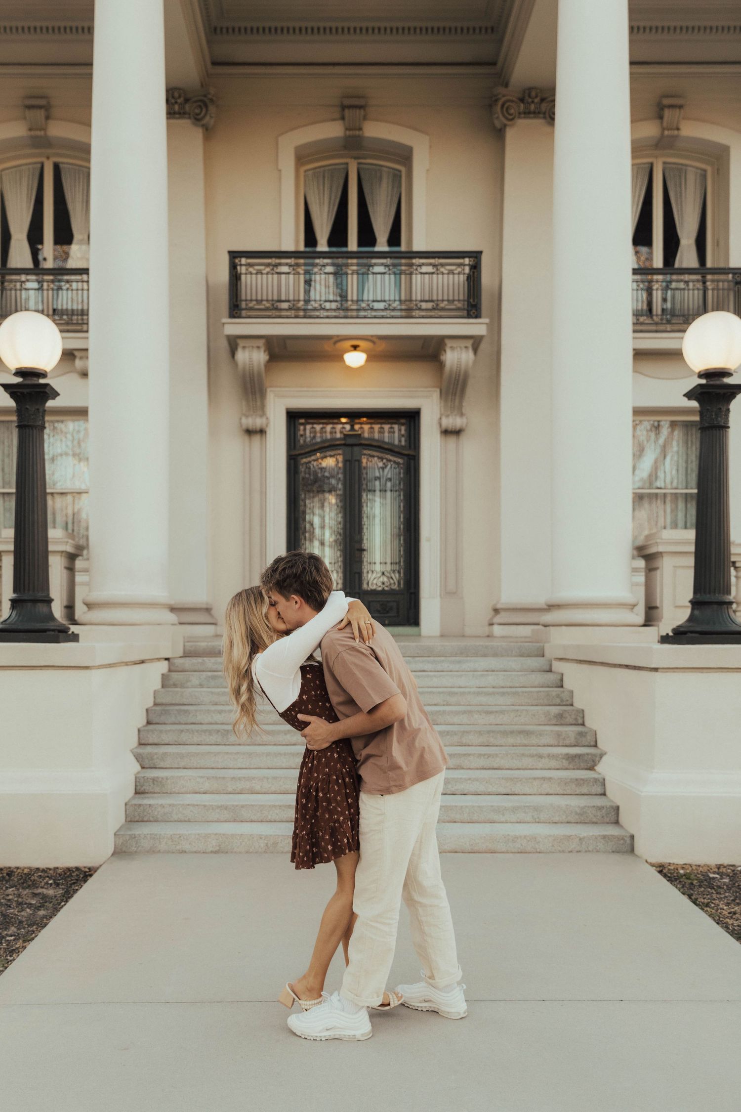 Romantic couple embraces in front of elegant historic building with columns and grand staircase at sunset.