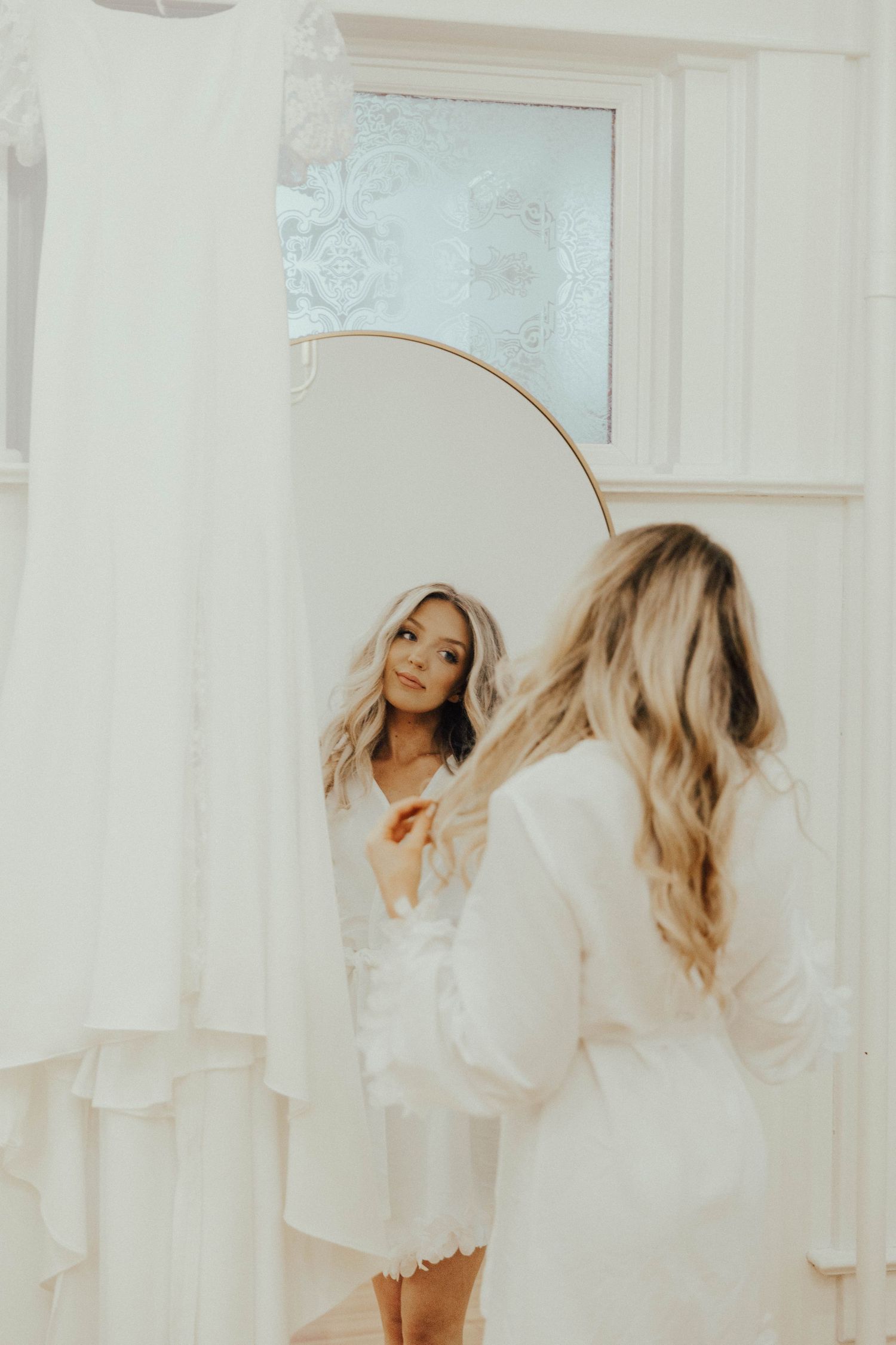 A reflection in a white-framed mirror shows someone in a light-colored outfit standing in an airy, bright room.