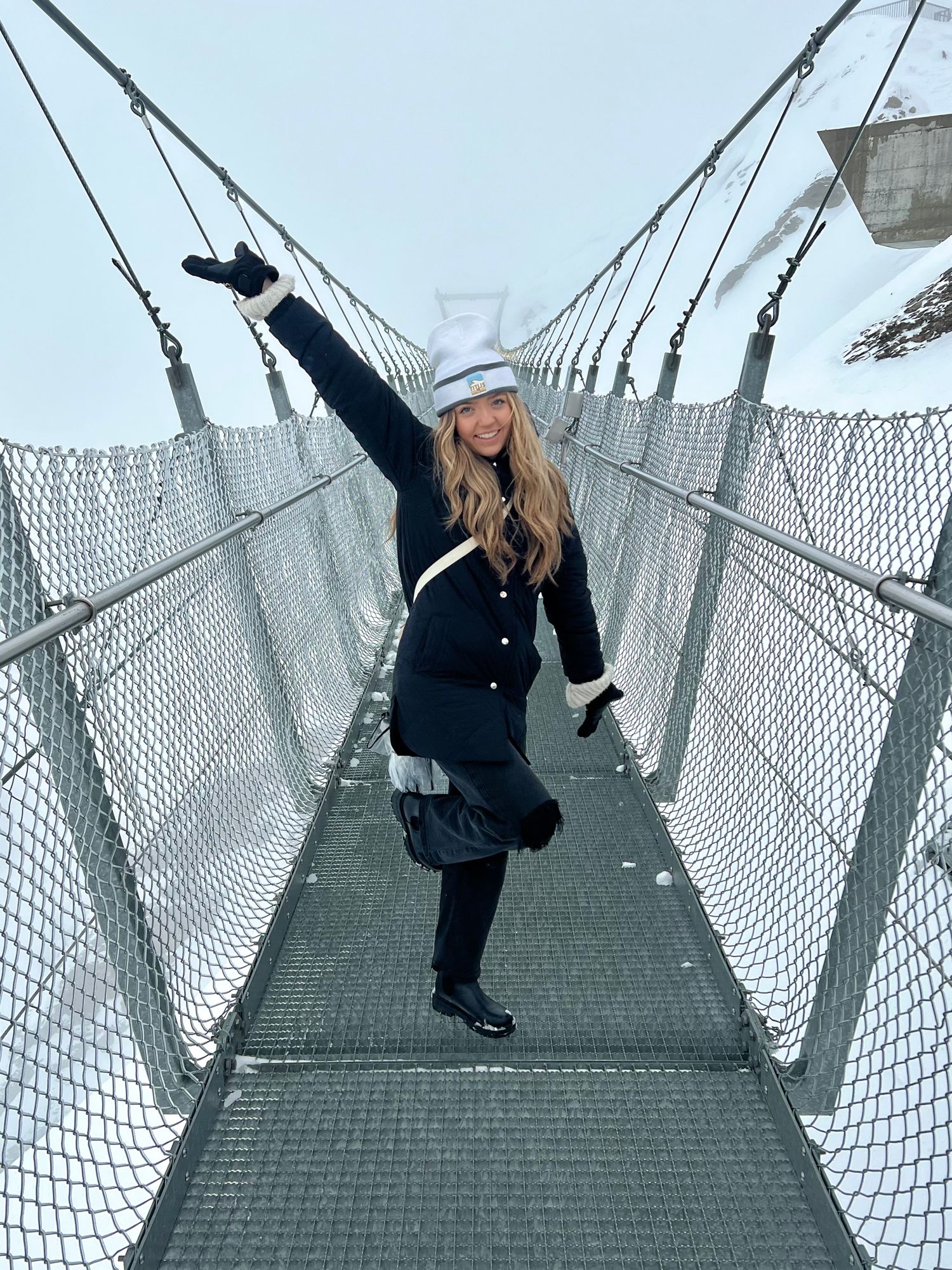 A person strikes a playful pose on a suspension bridge with safety netting, wearing winter clothing and a light hat.