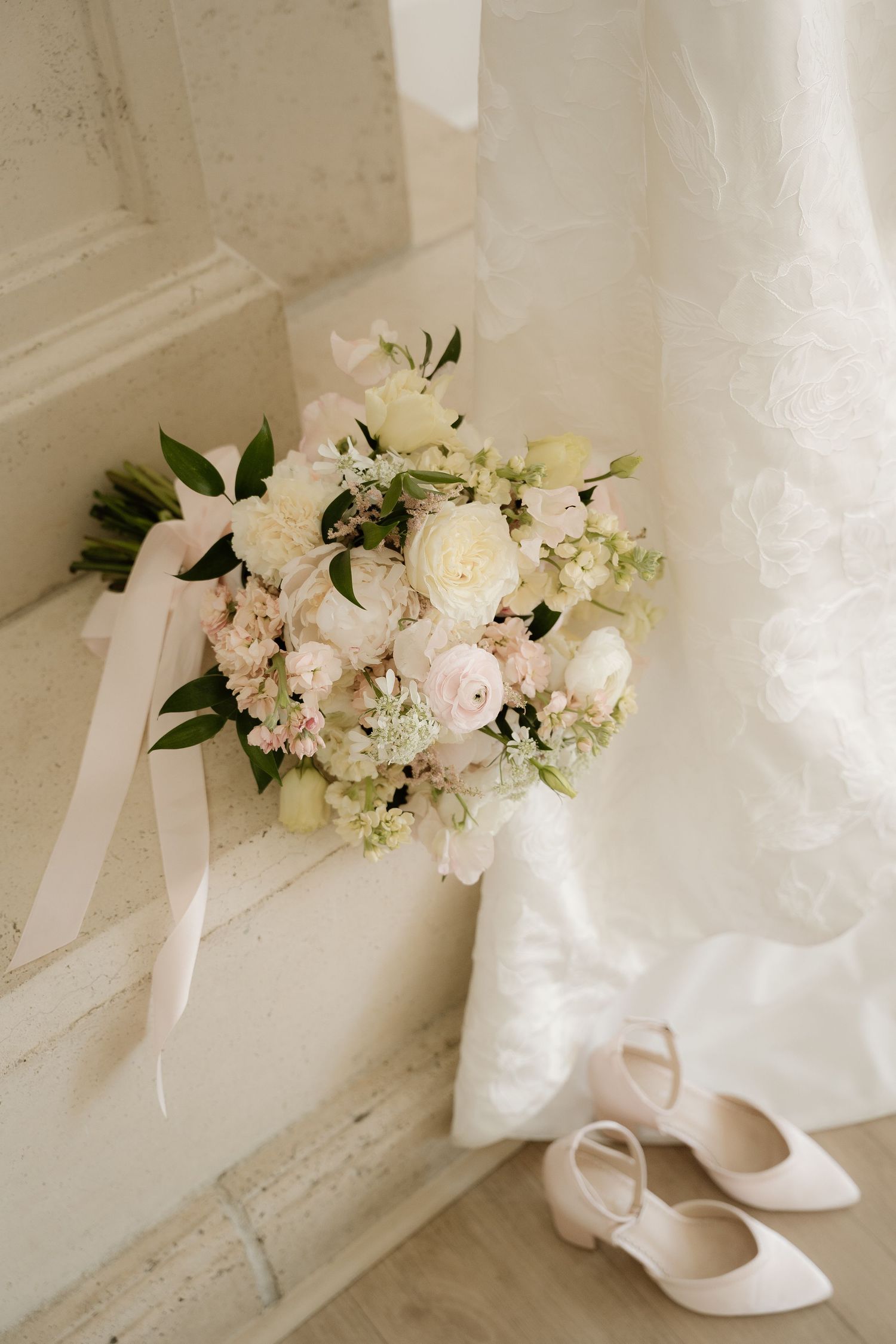 Elegant bridal bouquet of white and blush roses with ribbon beside ivory heels and lace wedding dress on marble steps.