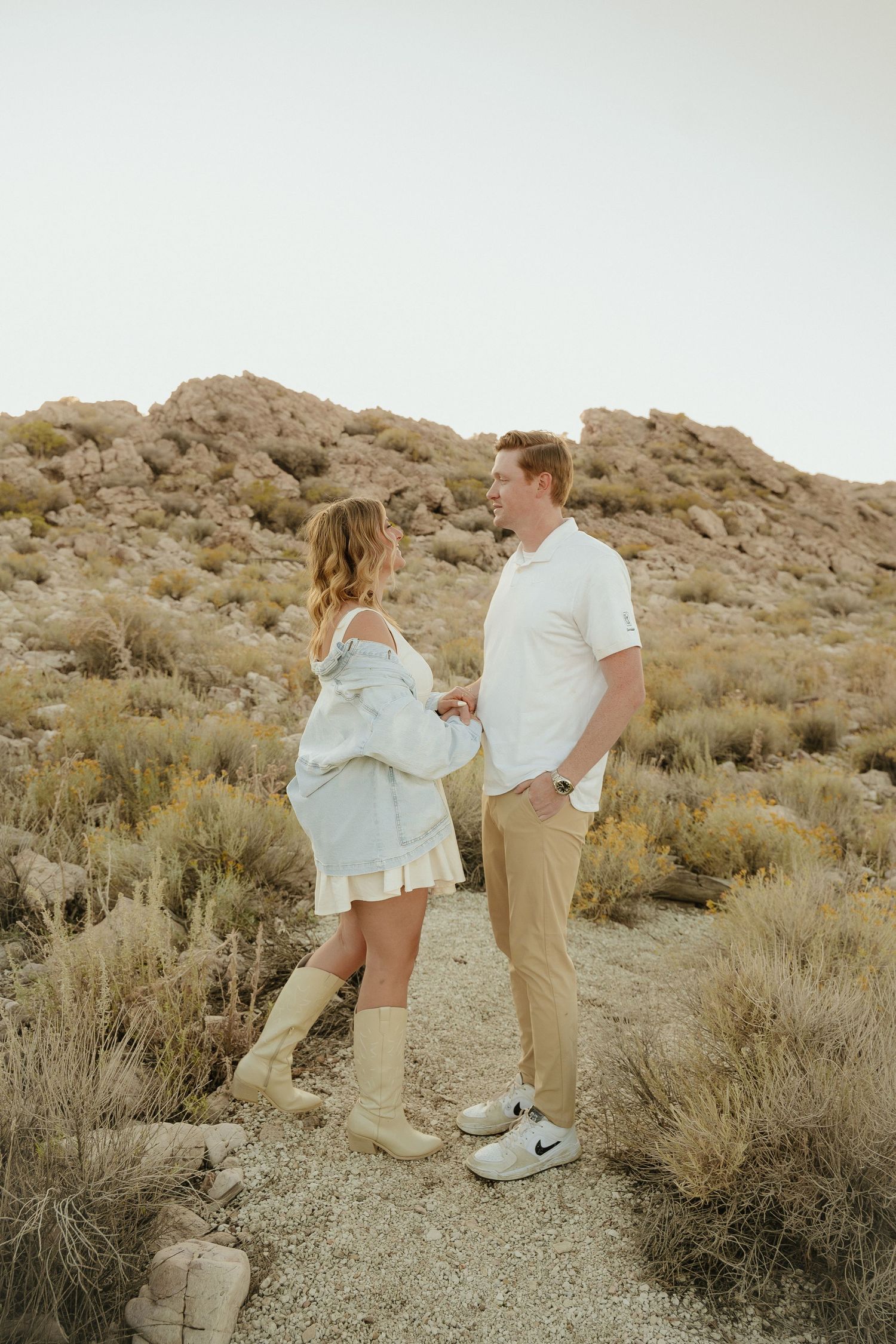 A couple holds hands on a desert trail surrounded by rocky hills and sagebrush, dressed in casual white and tan outfits.