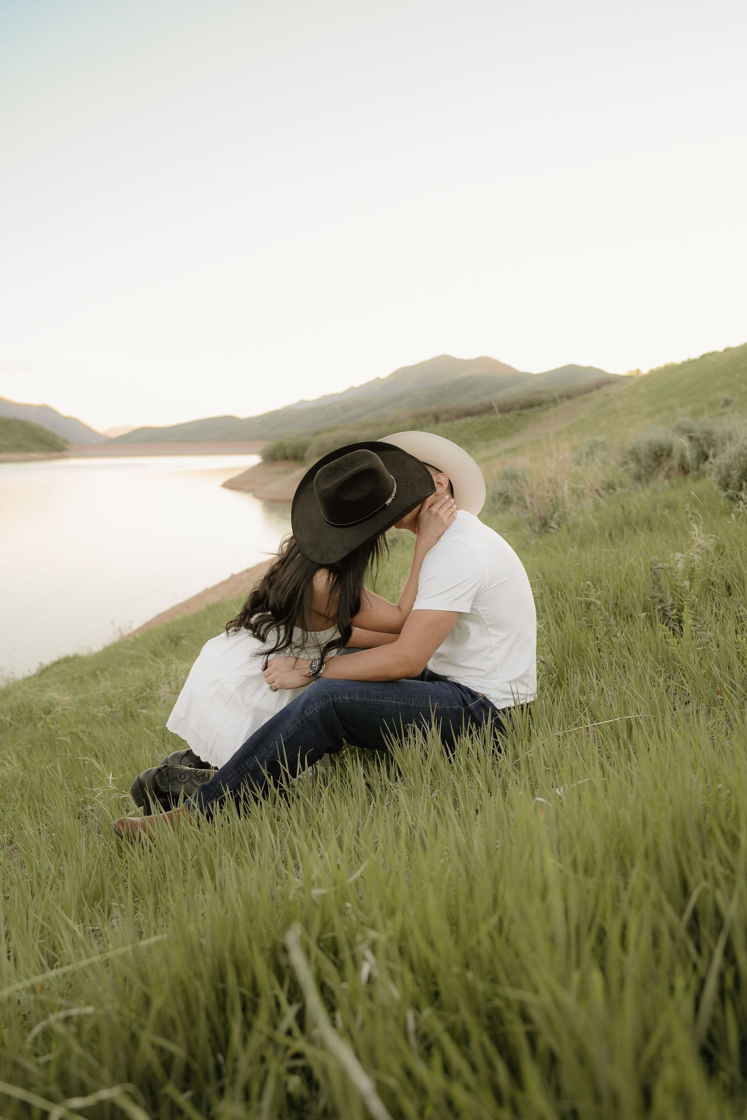 Couple sharing a romantic kiss on a grassy hillside by a lake at sunset, man in white tee and cowboy hat.