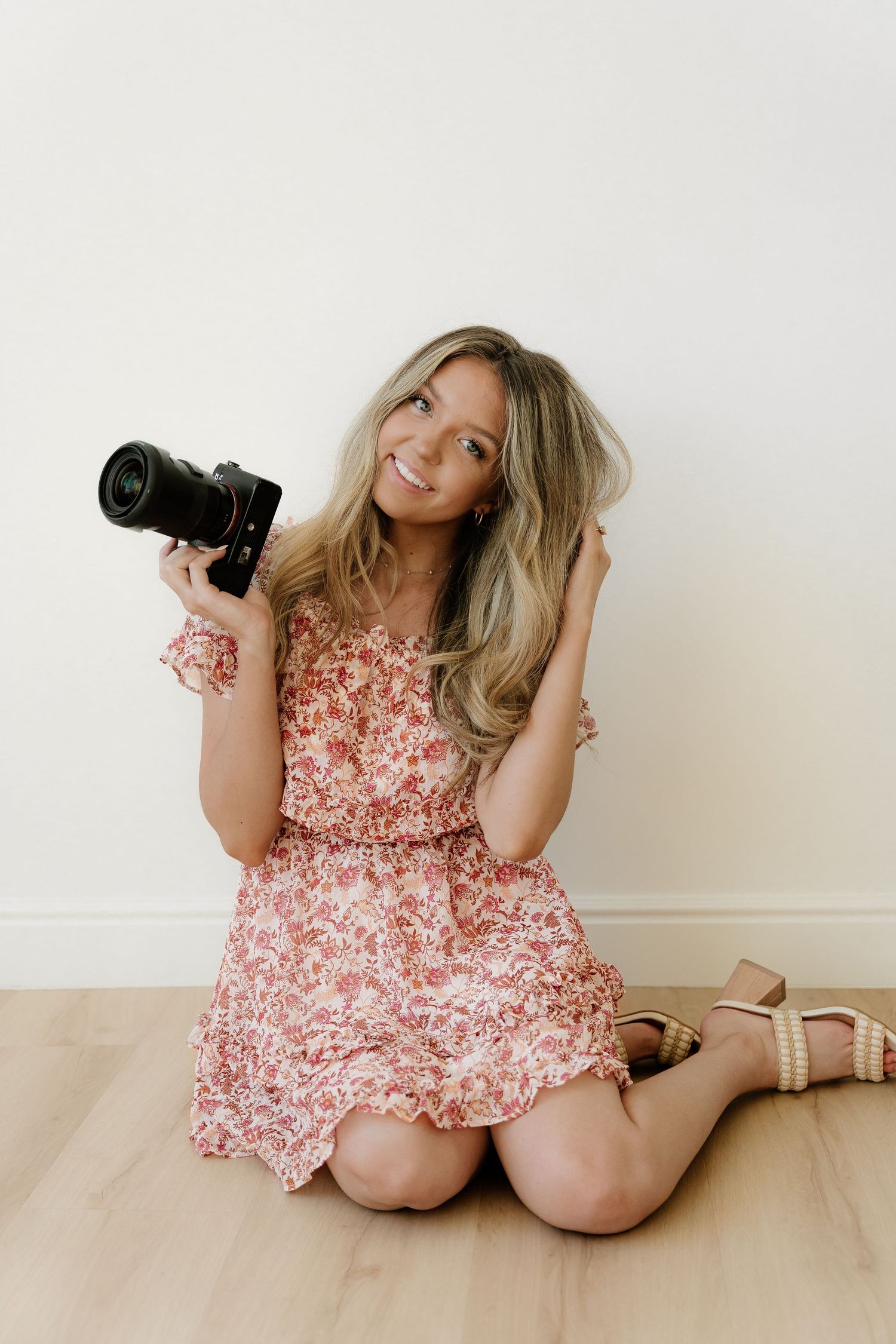 Photographer in floral pink dress sits on wooden floor holding professional camera while posing with blonde hair.