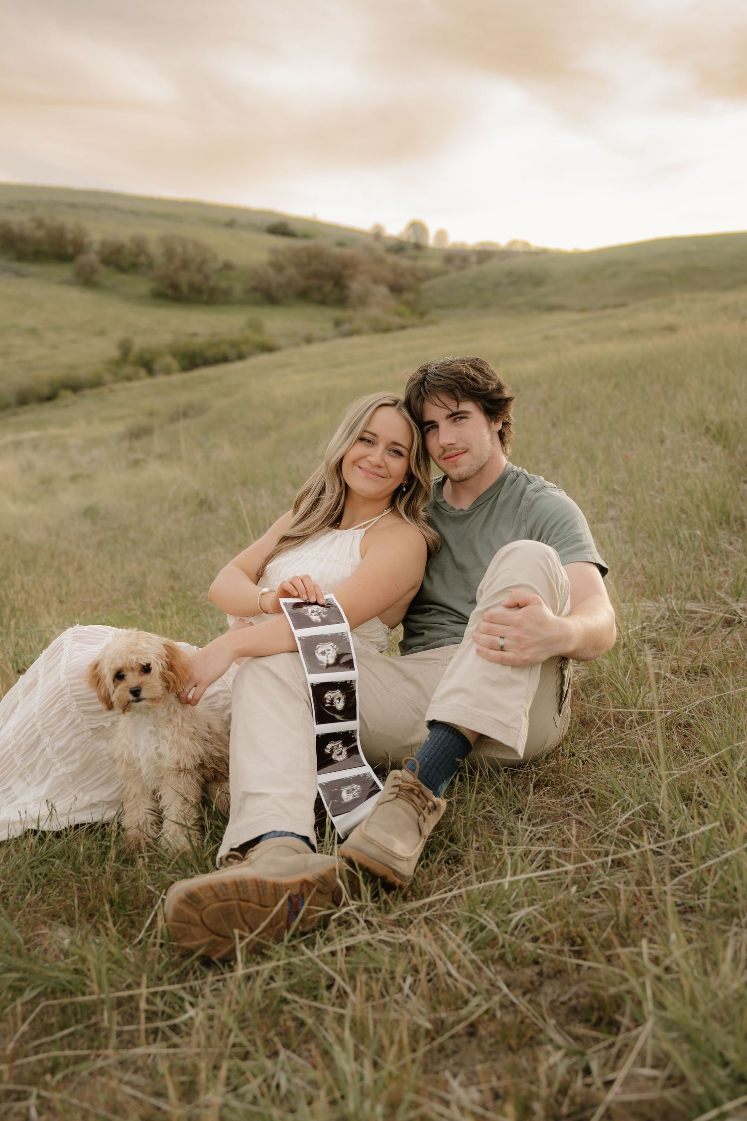 A couple sits on a grassy hill holding ultrasound photos with a small dog, announcing their pregnancy outdoors.