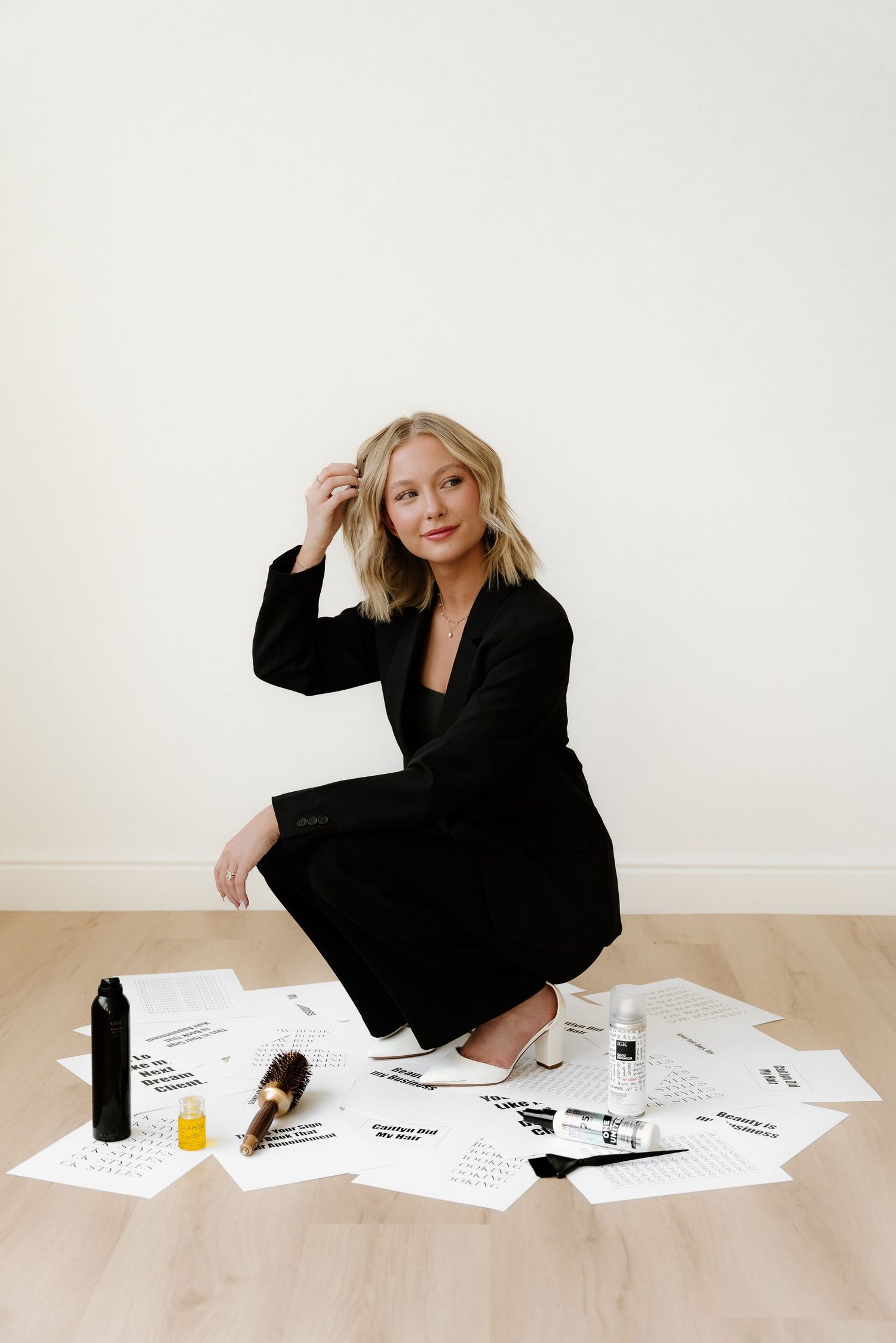 Professional sitting on floor in black dress surrounded by scattered papers with beauty care items on wooden surface.