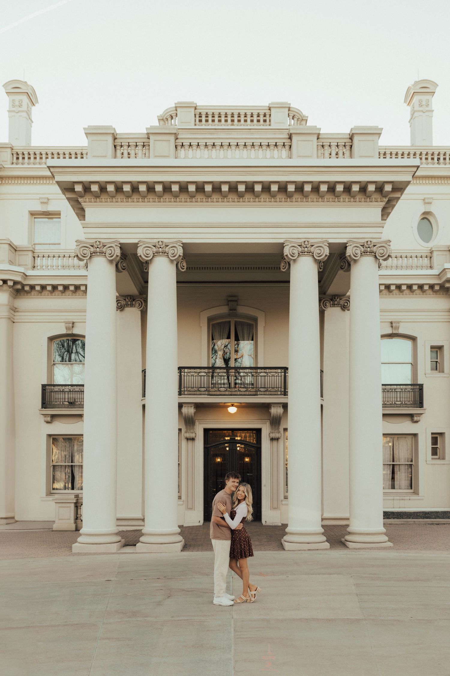 Two people embrace in front of a grand neoclassical building with towering white columns and ornate architectural details.