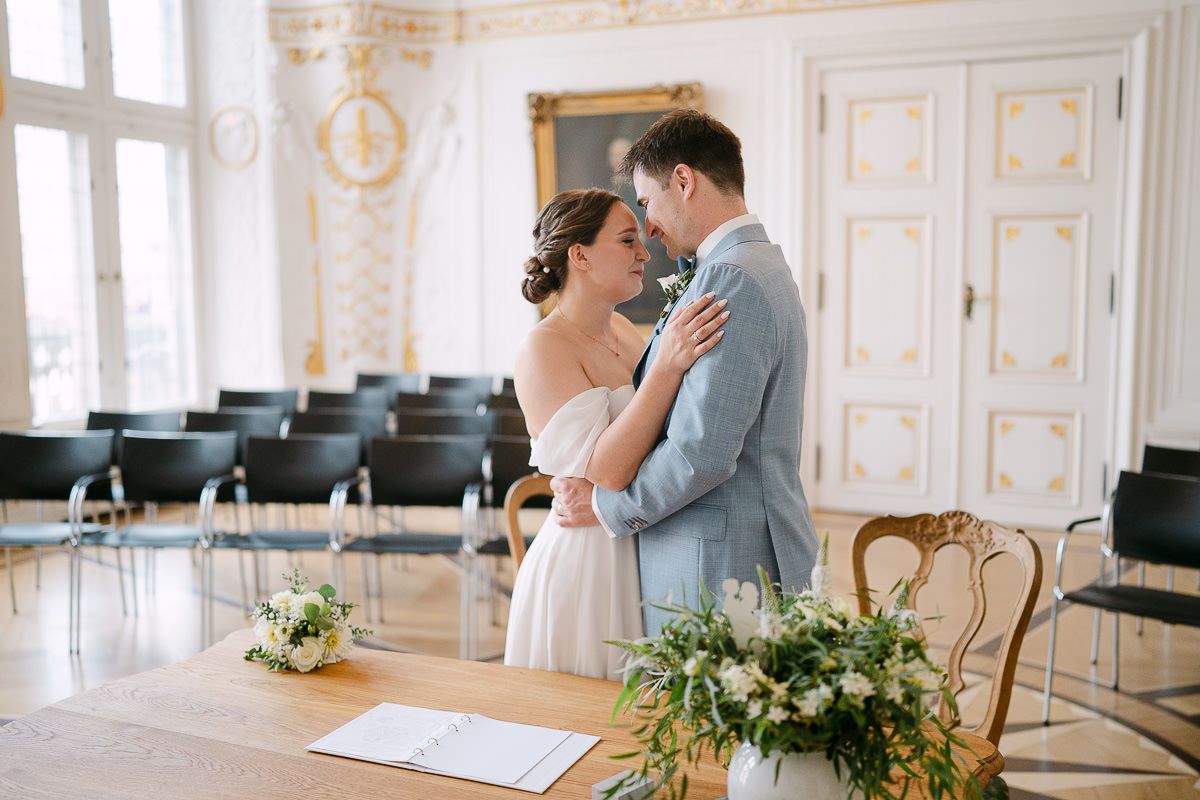 Ein Brautpaar genießt den zärtlichen Moment im eleganten weißen Saal mit kunstvollen architektonischen Details und Blumenarrangements.