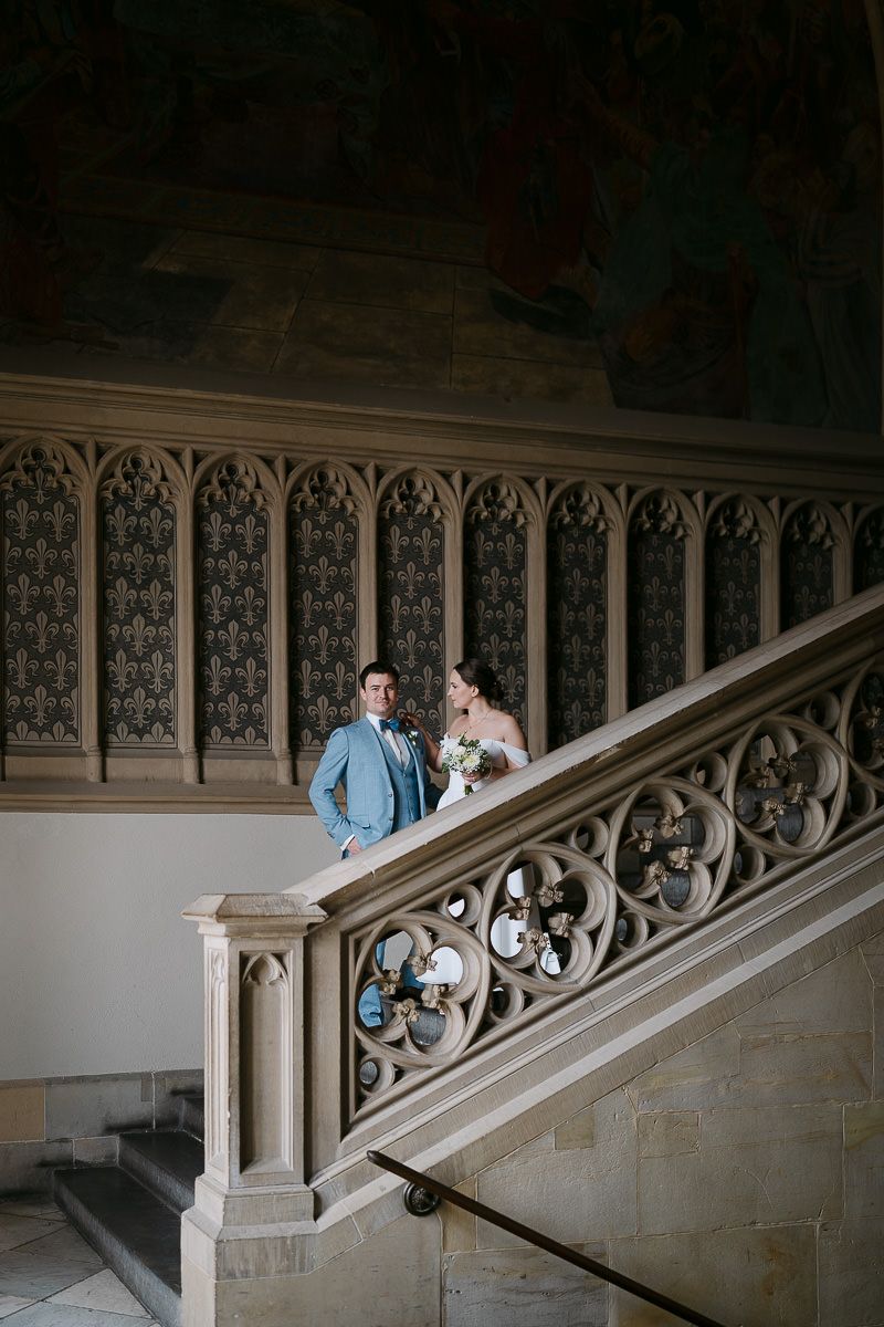 Ein Brautpaar steht gemeinsam auf einer reich verzierten Steintreppe mit dekorativem Geländer im eleganten Interieur des Aachener Rathauses.