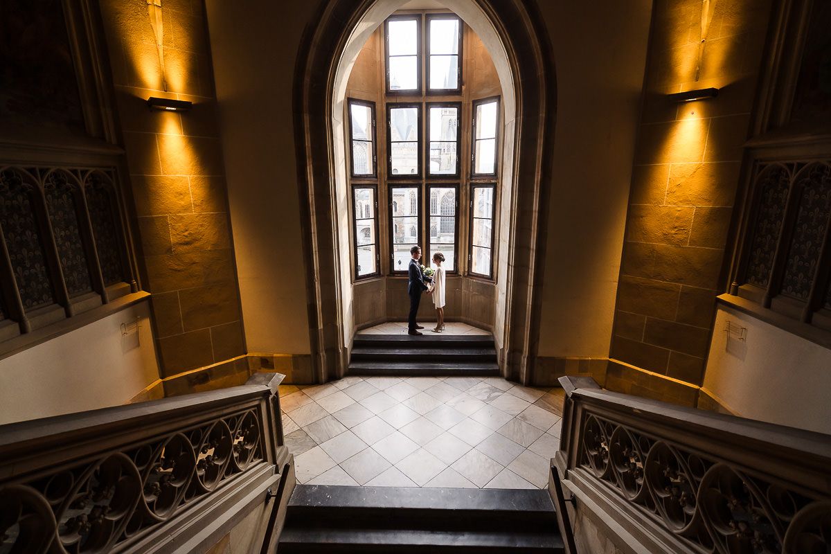 Sonnenlicht strömt durch gotische Fenster in das Treppenhaus des Rathauses Aachen mit einem Brautpaar im Panoramafenster.