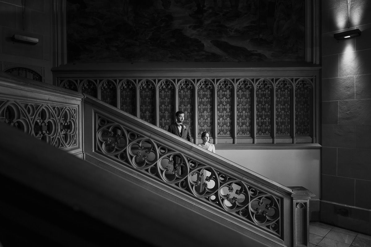 Eine eindrucksvolle Schwarz-Weiß-Fotografie der reich verzierten Treppe mit dekorativem Geländer, die von einer Wandleuchte beleuchtet wird.