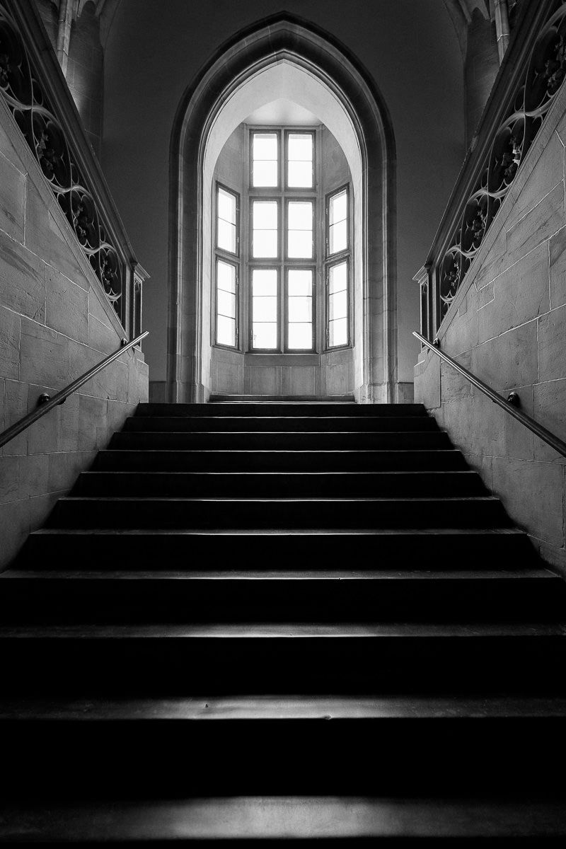 Schwarz-Weiß-Aufnahme einer imposanten Steintreppe, die zu einem gotischen Bogenfenster im Inneren des Rathauses Aachen führt.