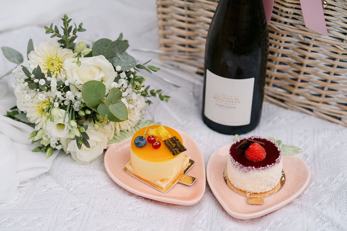 Picnic flatlay with champagne, pastries, and bouquet in park.