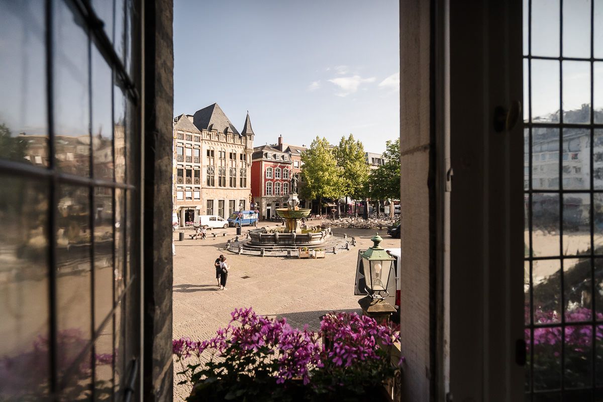 Durch große Fenster bietet sich ein Panoramablick auf einen historischen Marktplatz mit Gebäuden und farbenprächtigen Blumen im Vordergrund.