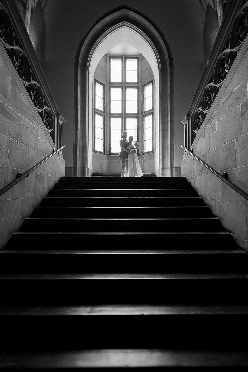 Schwarz-Weiß-Fotografie einer prunkvollen Treppe mit Bogenfenstern, die im historischen Rathaus Aachen dramatische Schatten werfen.