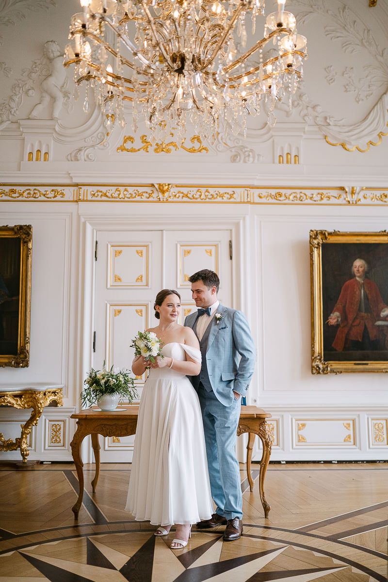 Ein elegantes Brautpaar steht im prunkvollen weißen Saal mit Kristallleuchter, vergoldeten Rahmen und barocker weiß-goldener Dekoration.