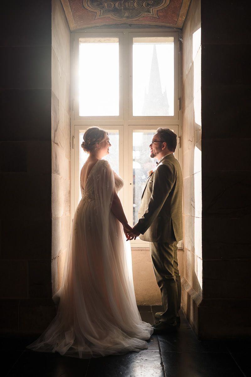 Ein im Gegenlicht stehendes Paar in Hochzeitskleidung steht an einem sonnenbeschienenen Fenster und erzeugt so eine romantische Atmosphäre.