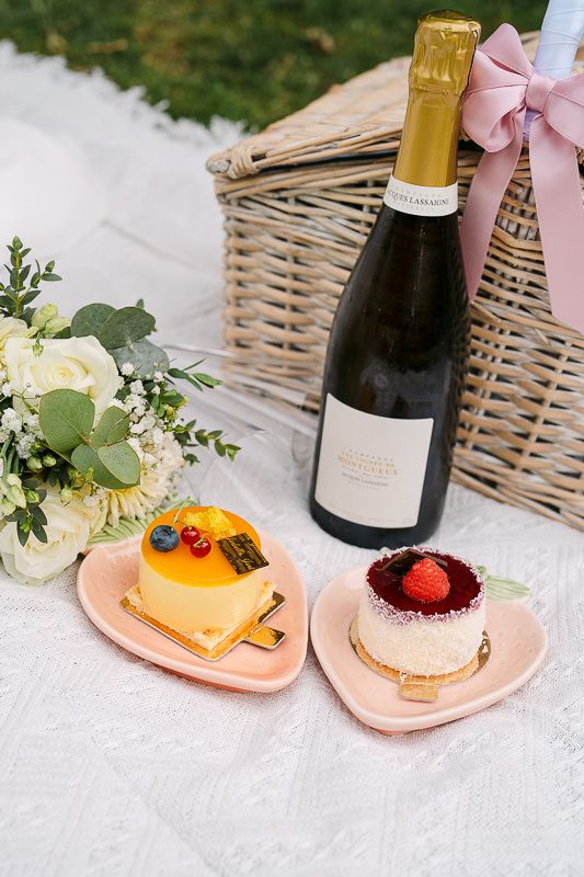 Picnic flatlay with champagne, pastries, and bouquet in park.