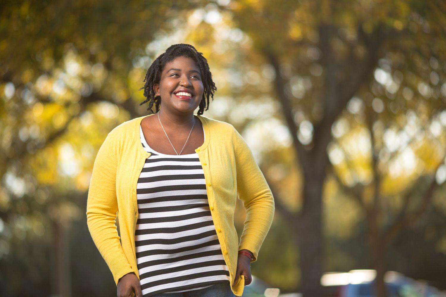 A joyful person in a yellow cardigan and striped shirt poses outdoors against an autumn background.