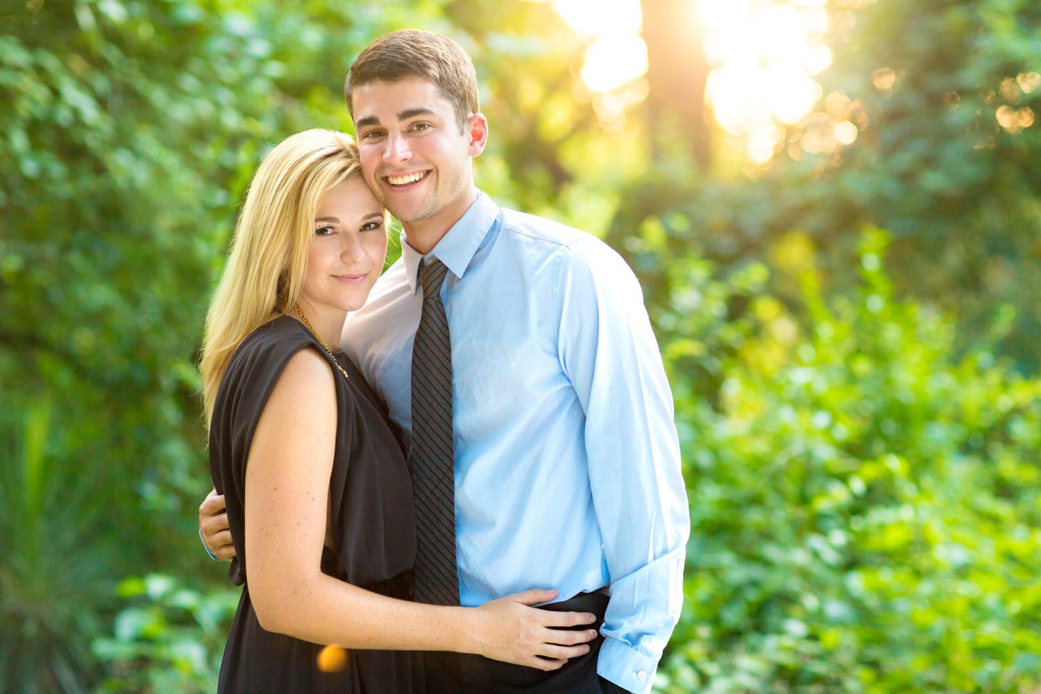 A couple embraces outdoors during golden hour with lush green foliage in the background.