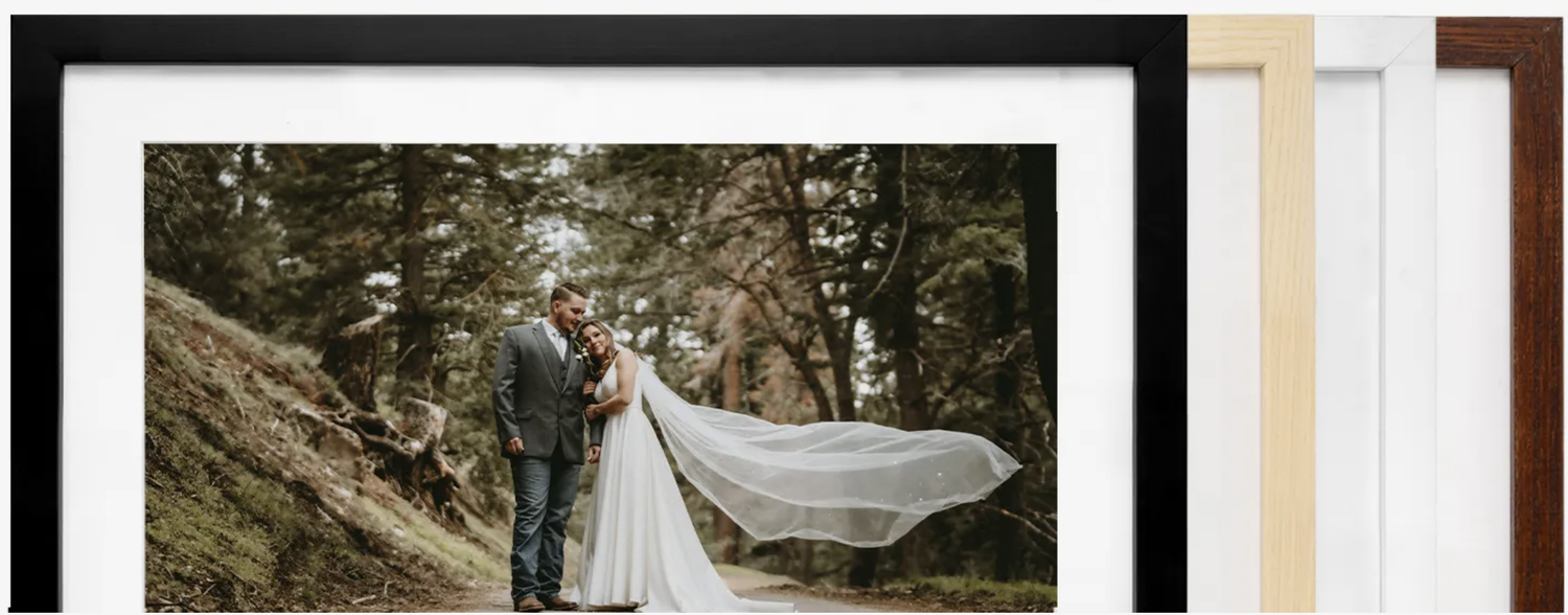 A couple in formal wedding attire embraces on a forest path as a long veil flows dramatically in the wind.