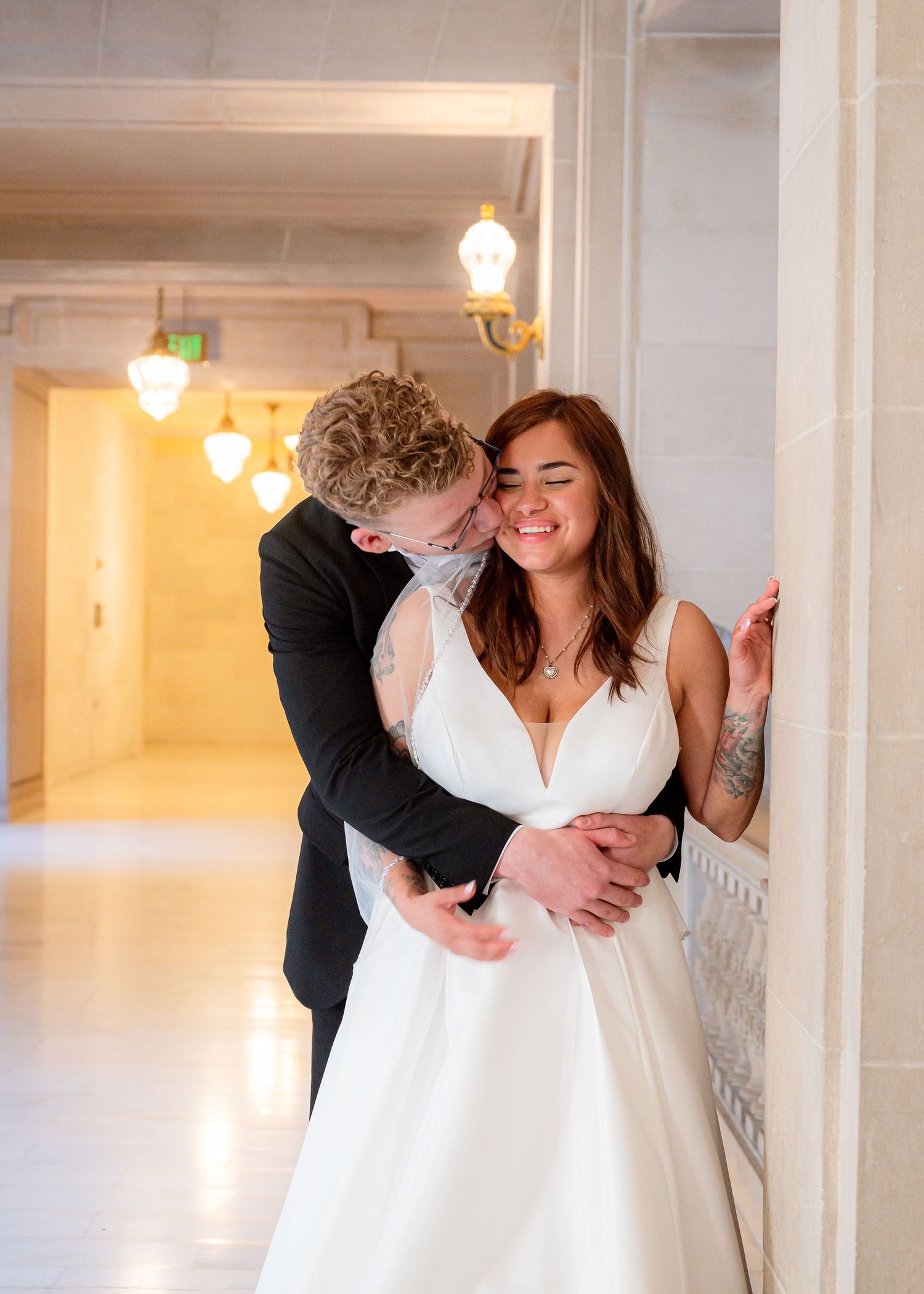A couple in white formal attire sharing romantic embraces in a bright hallway with pendant lights.