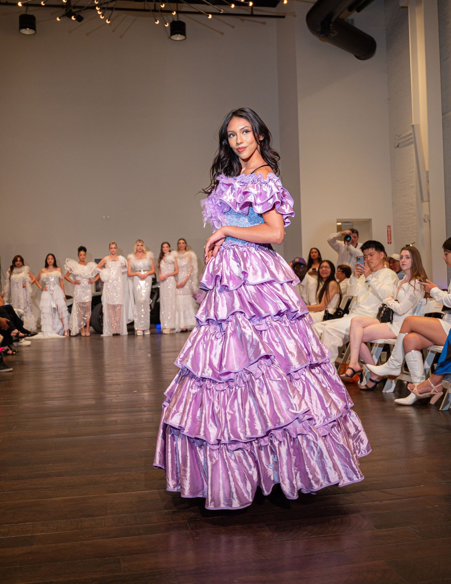 Fashion model walks runway in lavender multi-tiered ruffled ballgown during fashion show with audience in background.