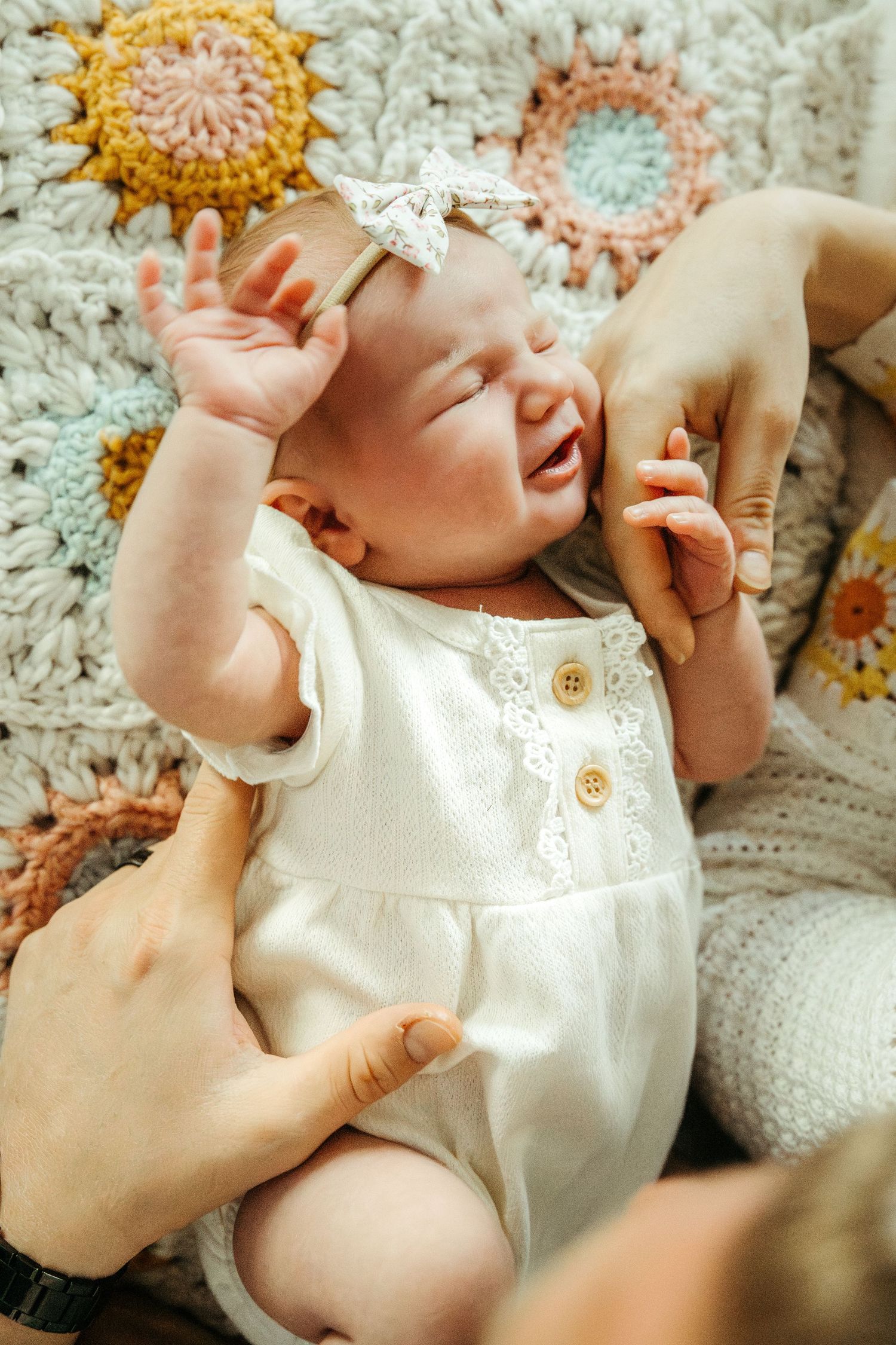 Hands tenderly dressing a baby in a white lace outfit against a crocheted blanket with colorful floral motifs.