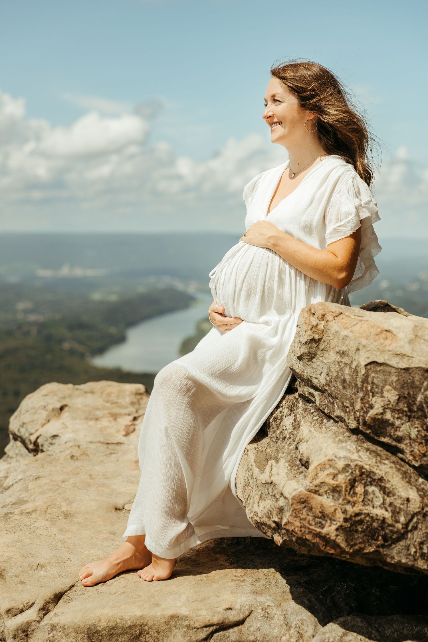 A person in a flowing white dress sits on a rocky cliff overlooking a scenic river valley on a sunny day.