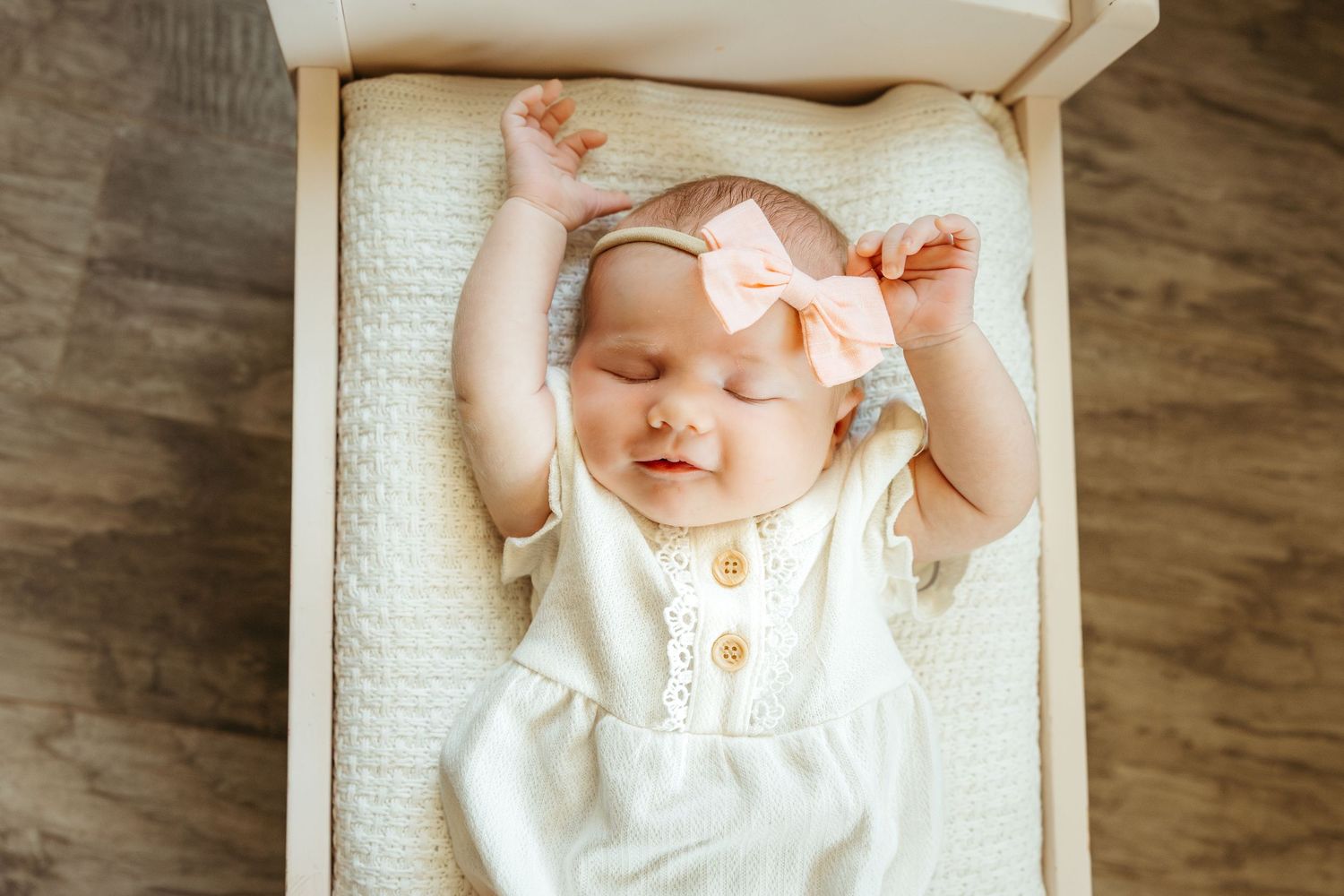 Sleeping baby wearing white outfit and peach bow rests peacefully on cream textured blanket.