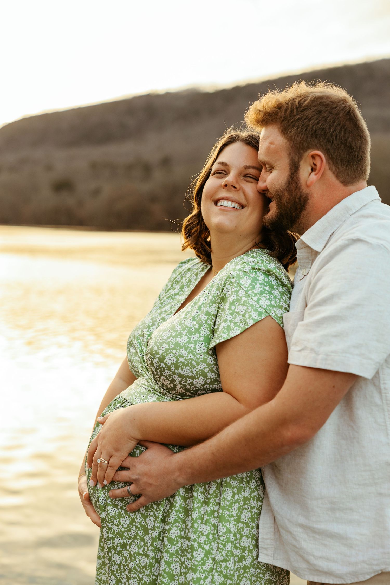 A couple embraces lovingly at sunset on a beach in a romantic maternity photoshoot.