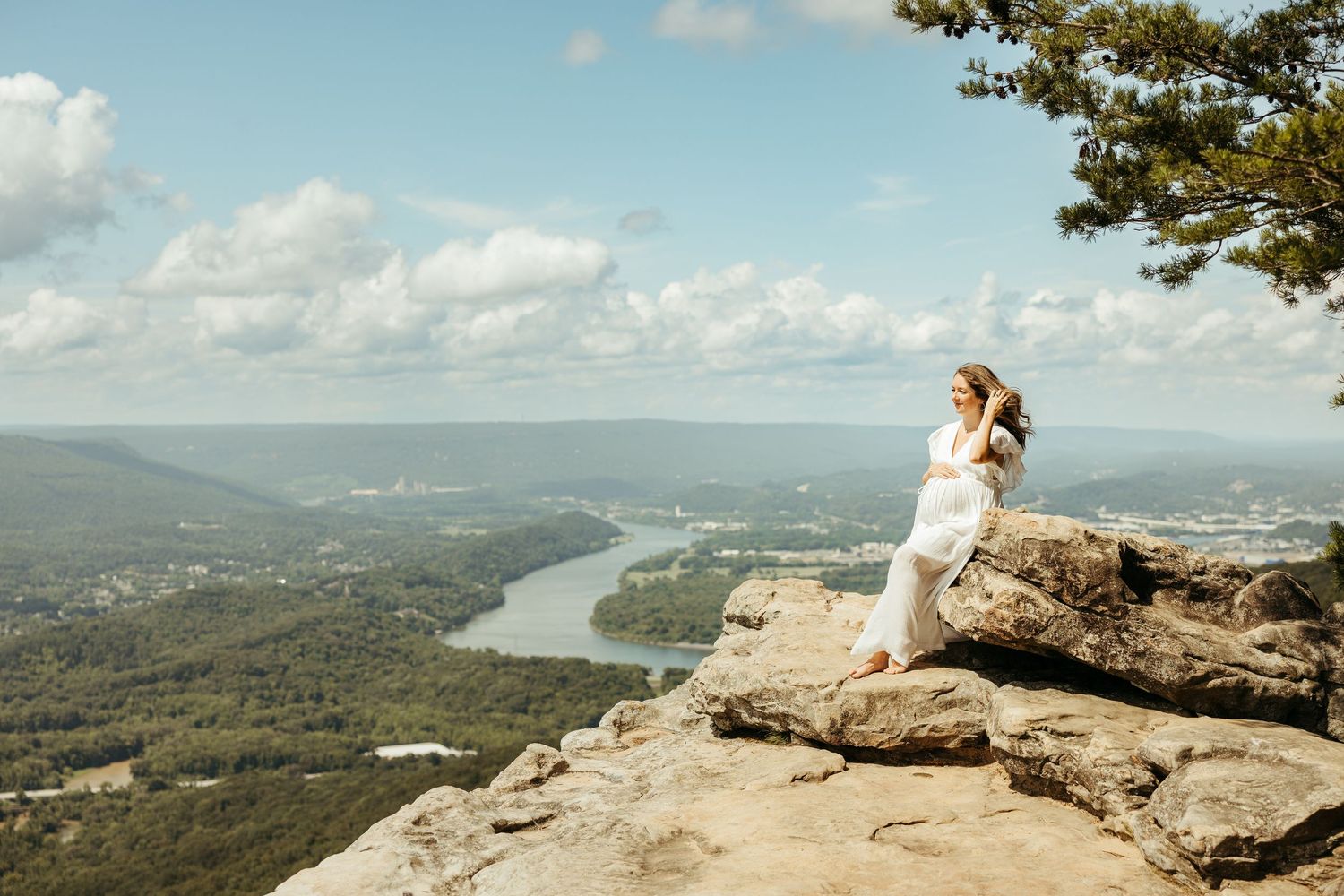 A scenic mountain overlook offers panoramic views of a lake and rolling hills on a sunny day with white clouds.