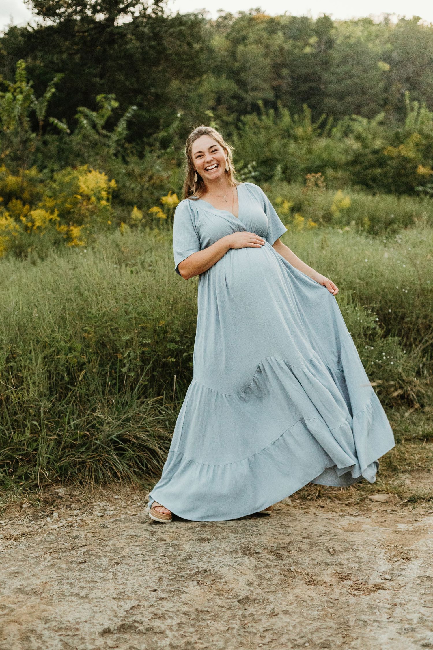 A light blue flowy maxi dress with tiered ruffles is showcased in a natural outdoor setting with greenery and sunlight.