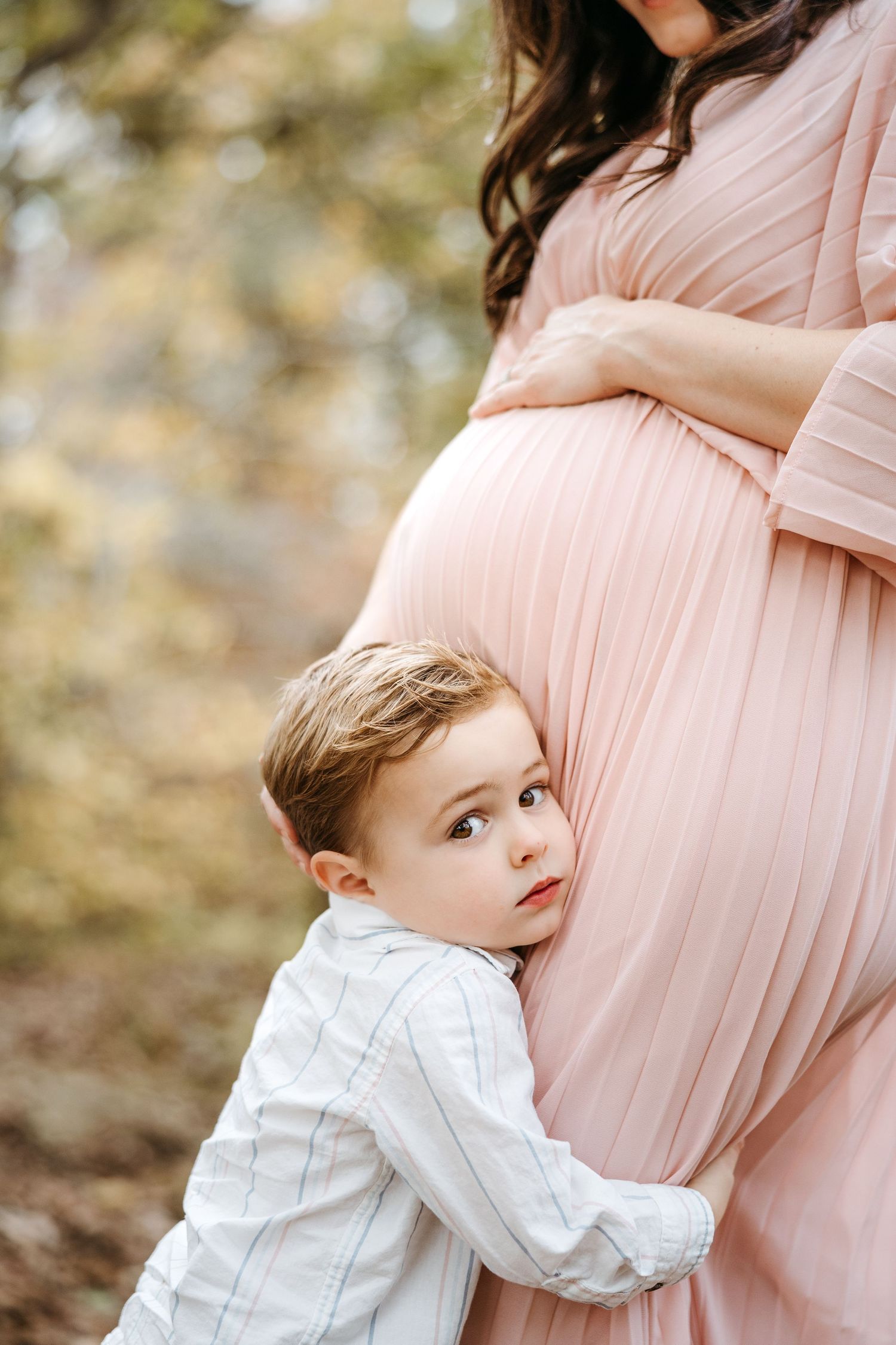 Young child in white shirt hugs pregnant woman wearing flowing blush pink maternity dress in outdoor setting.