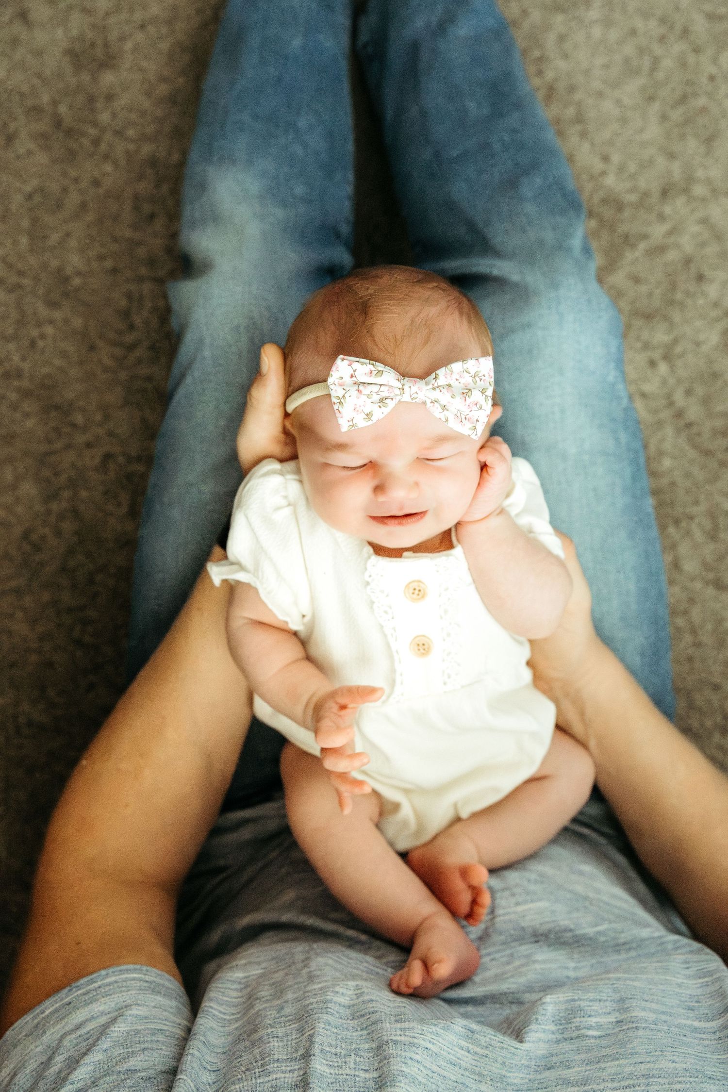 A baby in a white dress rests on someone's denim-clad lap.