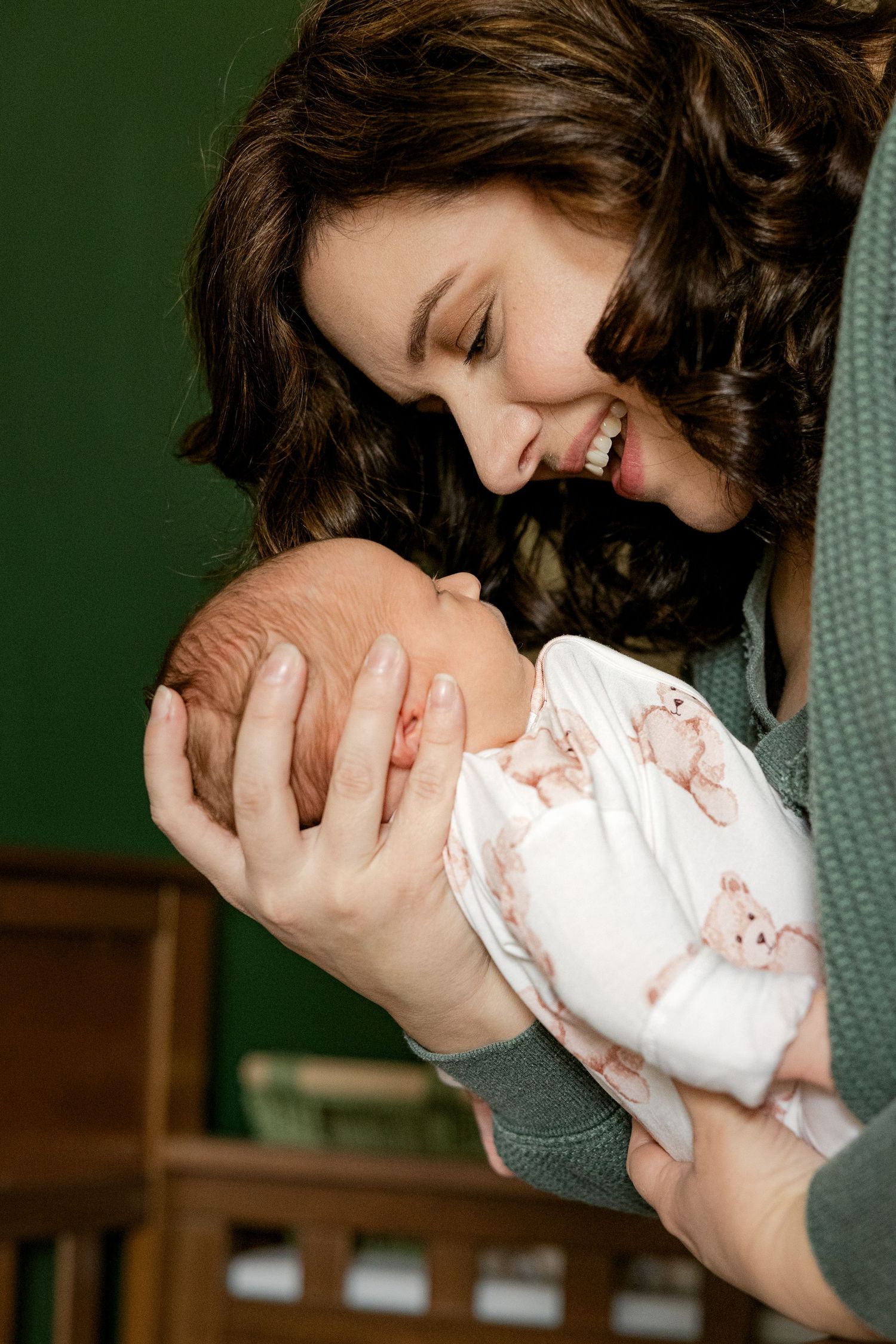 A woman with dark curly hair holds a newborn baby while smiling joyfully against a green background.