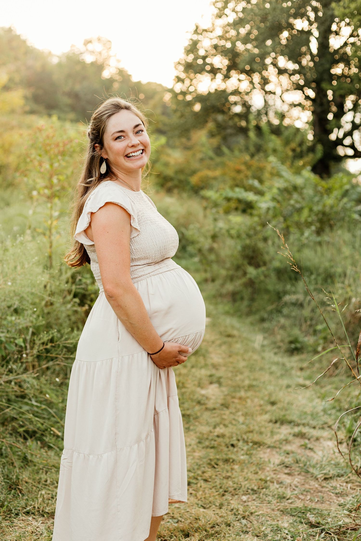 A maternity photoshoot in a natural outdoor setting with soft golden lighting and greenery in the background.