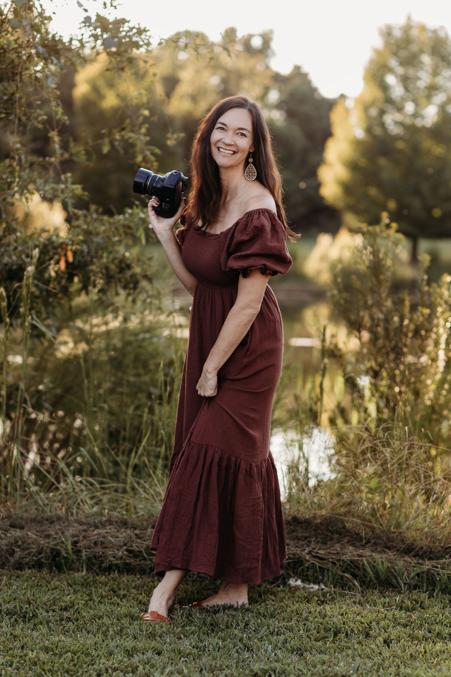 Smiling woman in burgundy off-shoulder dress holding camera outdoors near pond with lush greenery in golden hour light.